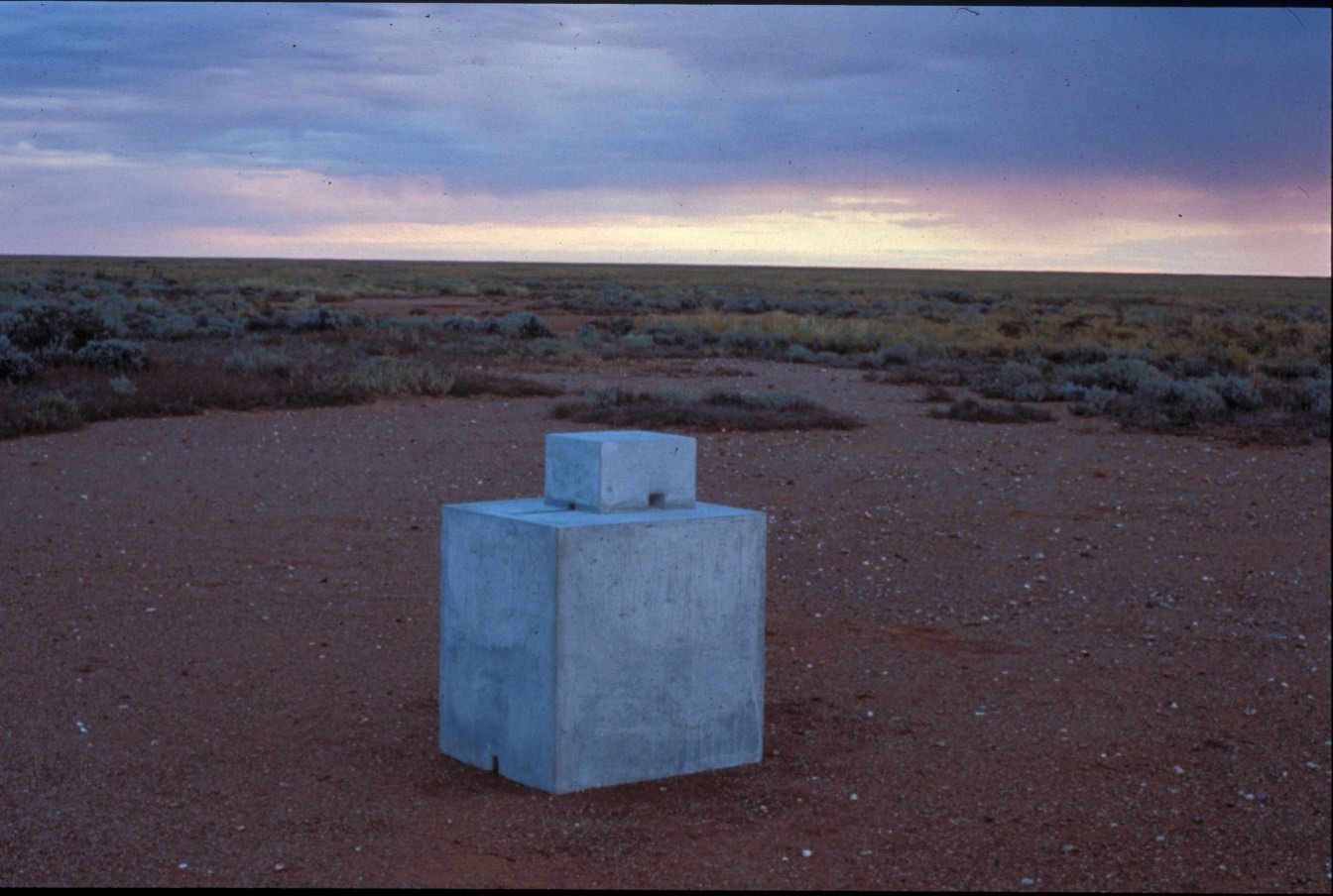 Sculpture. Two concrete blocks sit one atop the other in an expanse of absolutely flat desert. The top block is significantly smaller than the one below. The area around the blocks is sandy. A short distance away, a sea of scrubby green bush extends to the horizon.