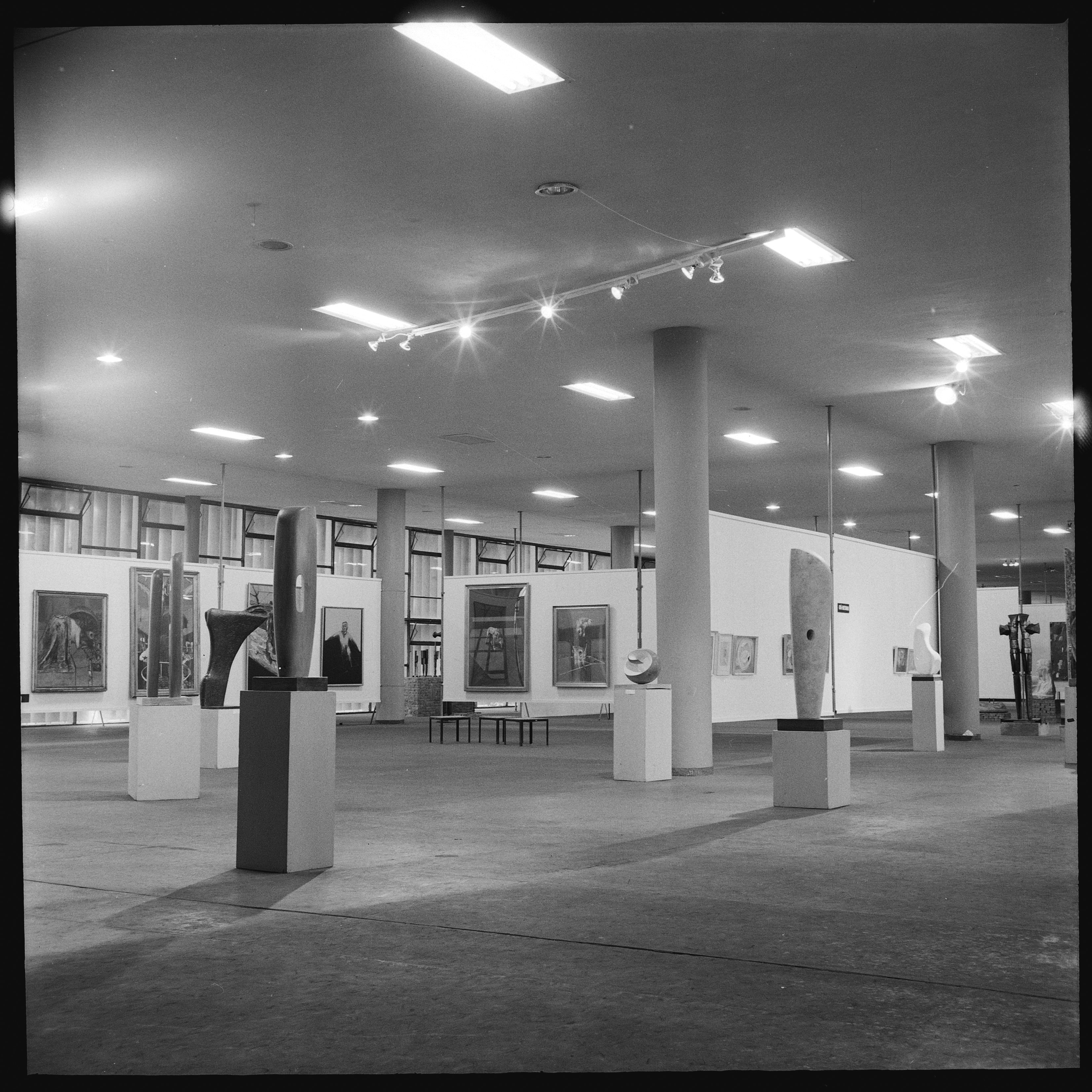 Installation photograph. Black and white photograph shows abstract sculptures on plinths arranged in a window-lined, open space. Works of art are hung on partial white walls.