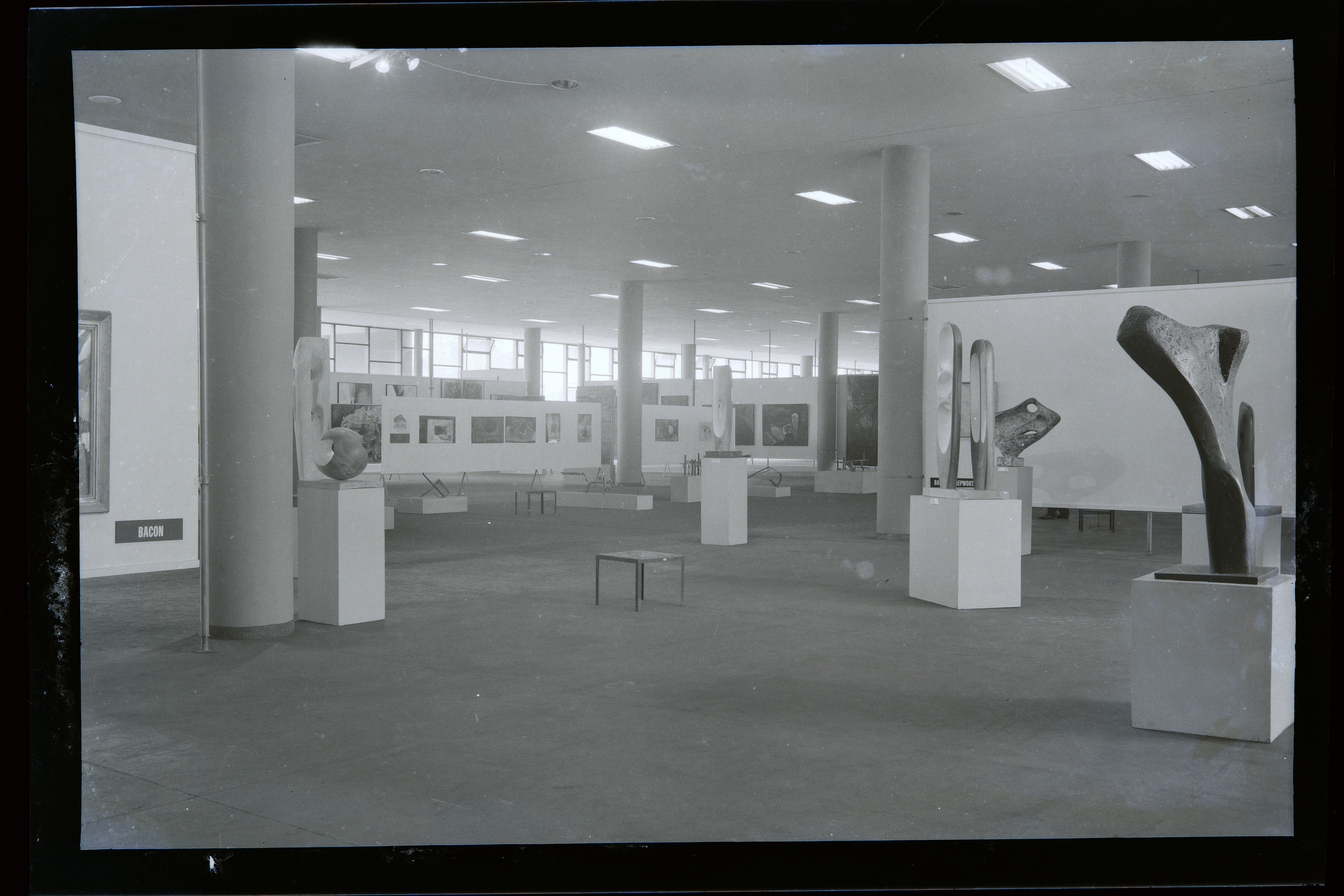 Installation photograph. Black and white photograph shows a window-lined, open space. Abstract sculptures on plinths are arranged throughout the space. In the far corner, works of art are hung on partial white walls.
