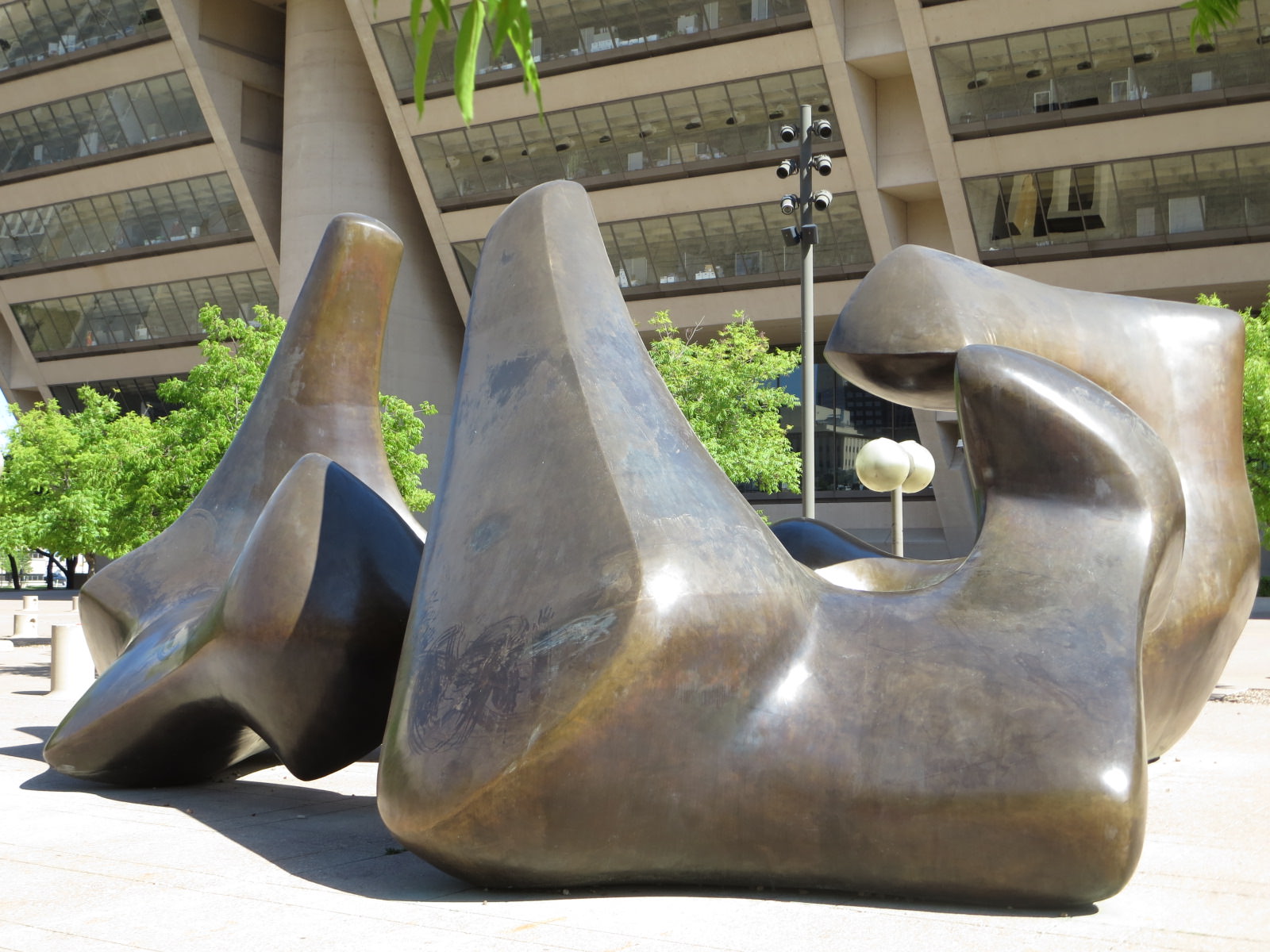 Sculpture. Three rounded, bronze forms sit in front of a building with a façade that angles out towards us. The bronze piece closest to us is roughly U shaped. The others rise and burble like a lava lamp.