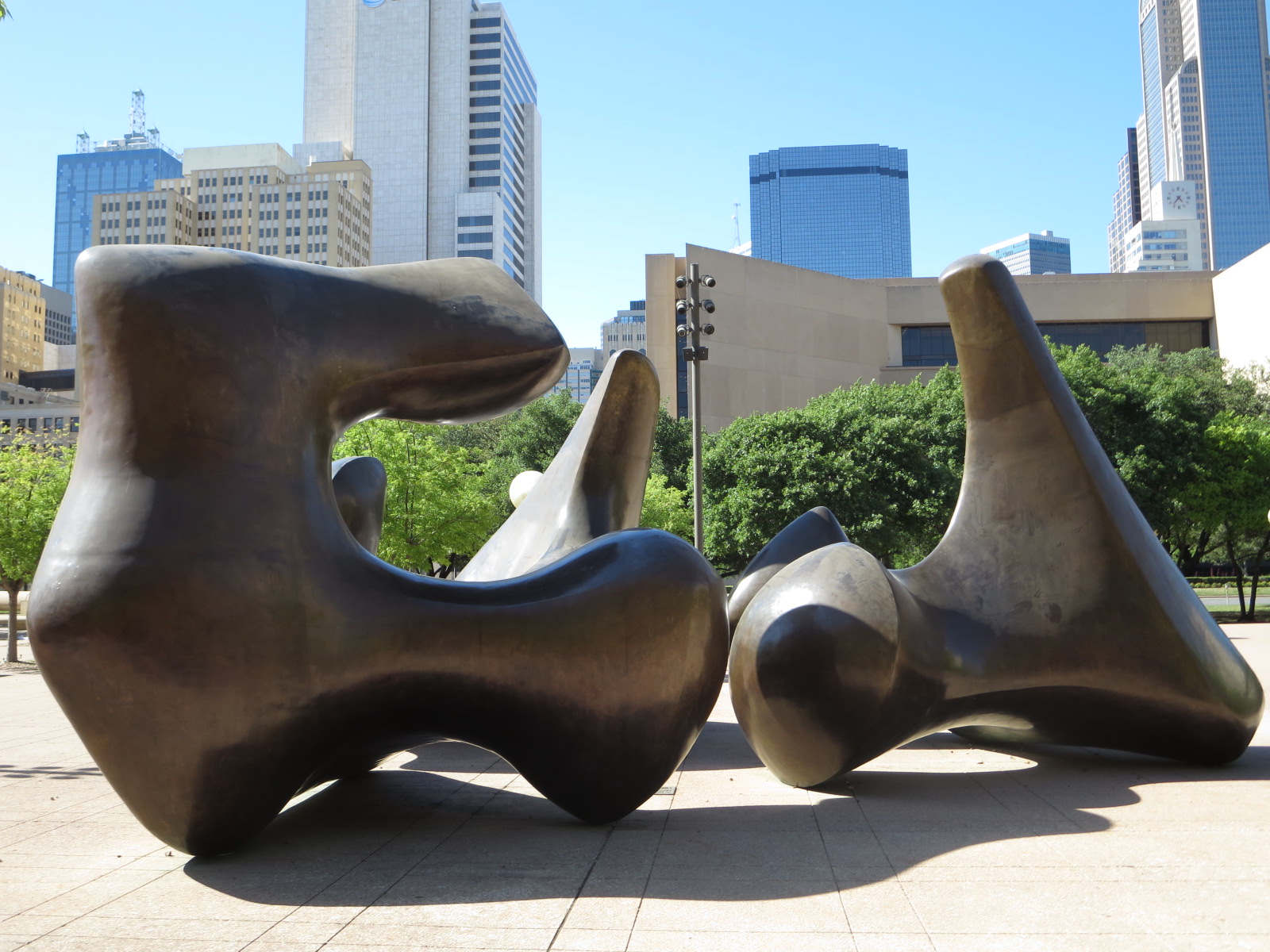 Sculpture. Three rounded bronze form face each other on a stone courtyard in front a city skyline. The forms are shaped like abstracted, vastly oversized vertebrae have straight backs and short, protruding arms with knobs at the end.