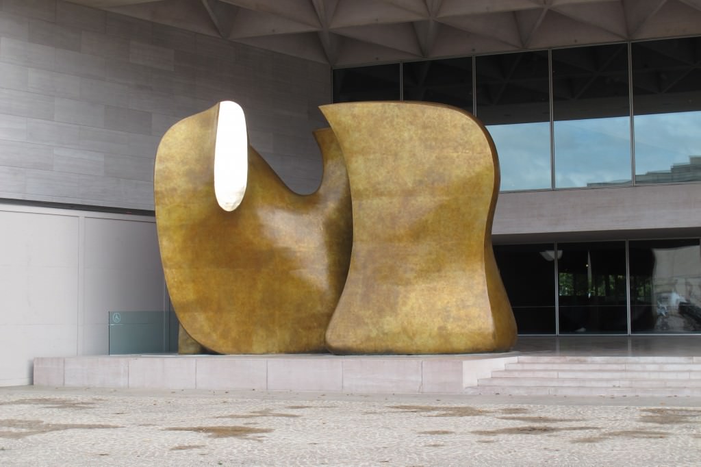 Sculpture. Two rounded bronze forms fill a two-storey, covered entryway. Most of the surface is burnished gold. An oval gleams brightly where the left-hand form seems to have been sliced.