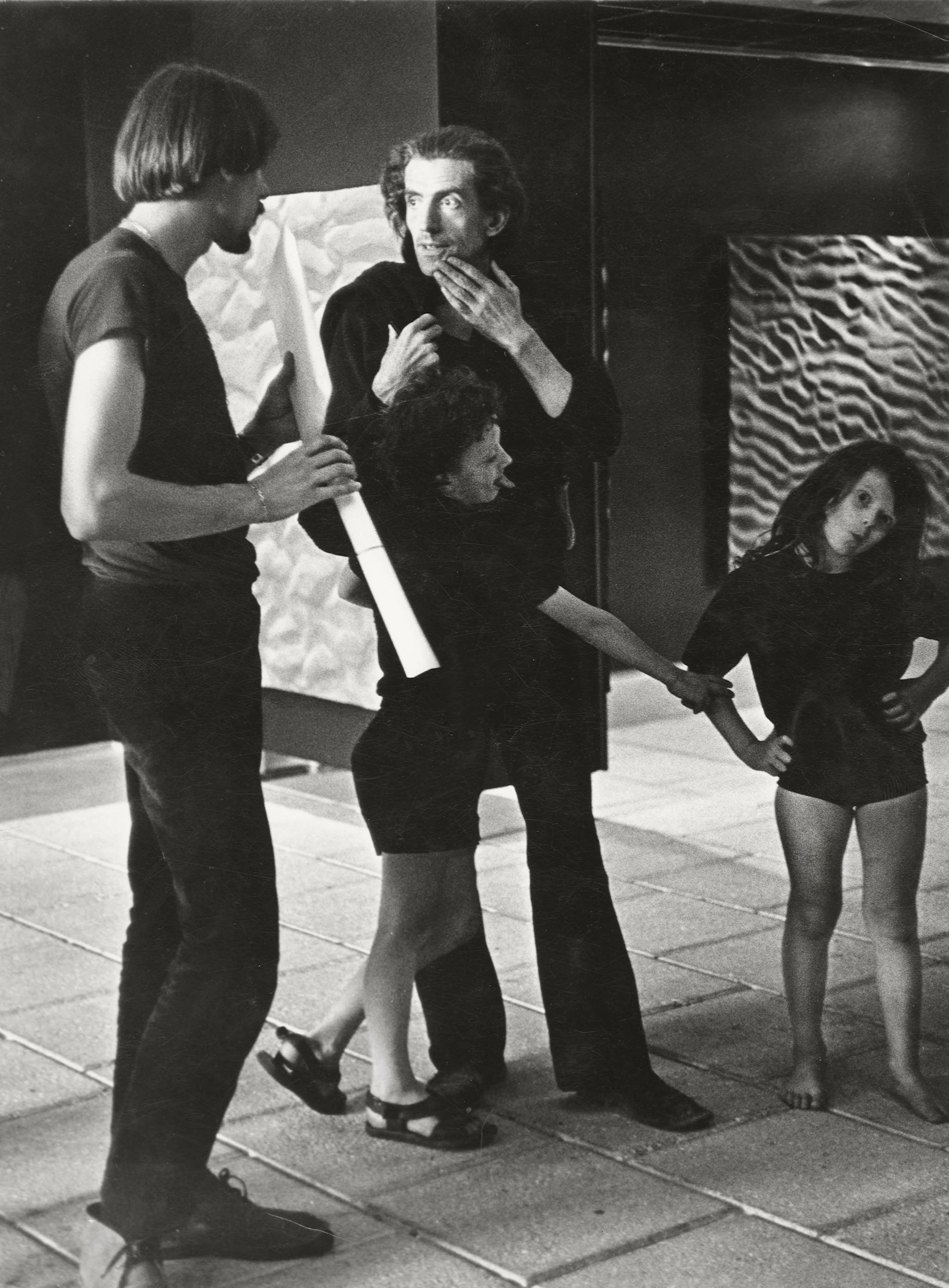 Black and white photograph. Two men and two children stand in an exhibition space. They all wear black and have pale skin.