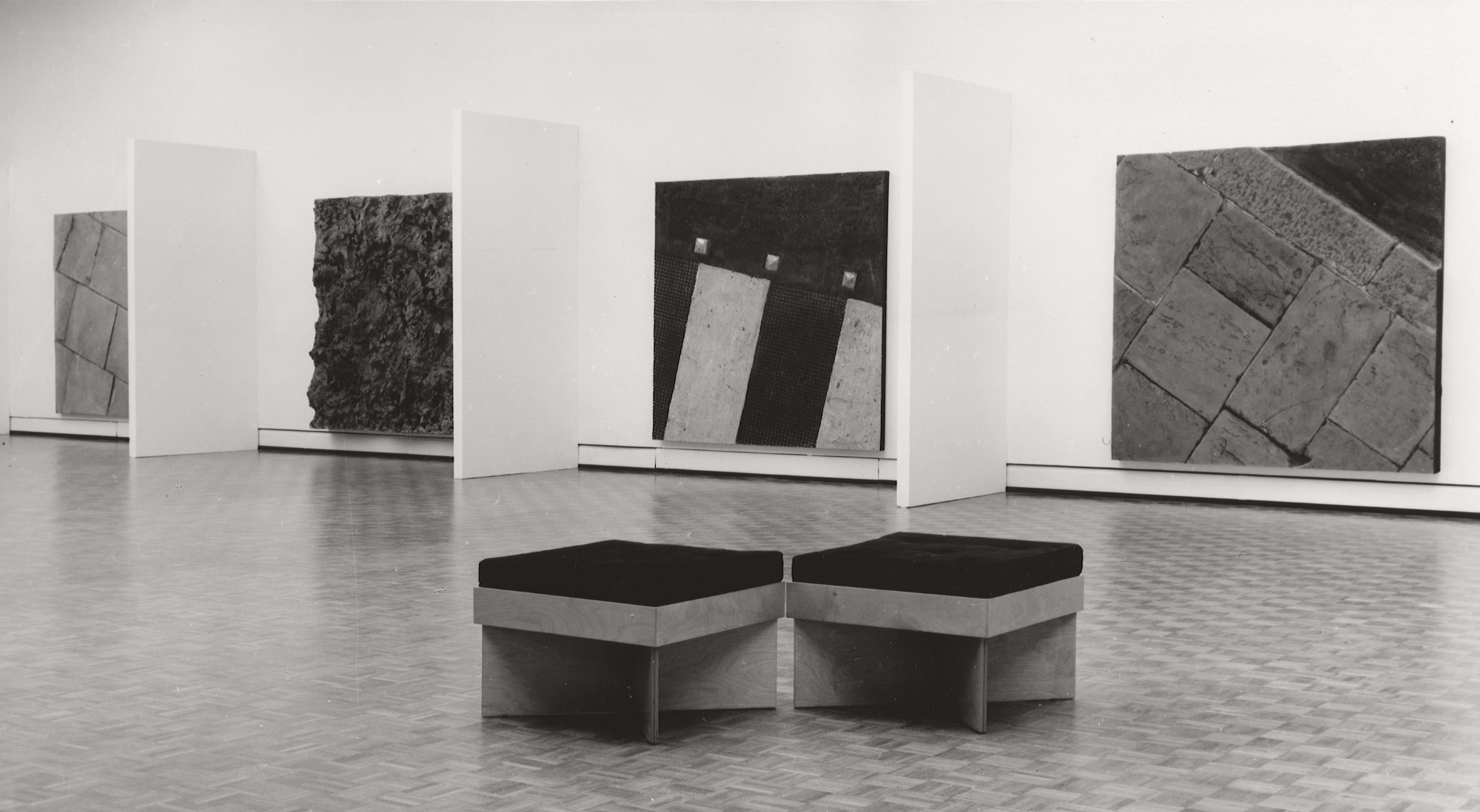 Installation photograph. Black and white photograph of four panels hung on a gallery wall. Each is square and is a cast of different kinds of ground, like pavers or a patch of dirt. Short projecting walls create barriers between them.