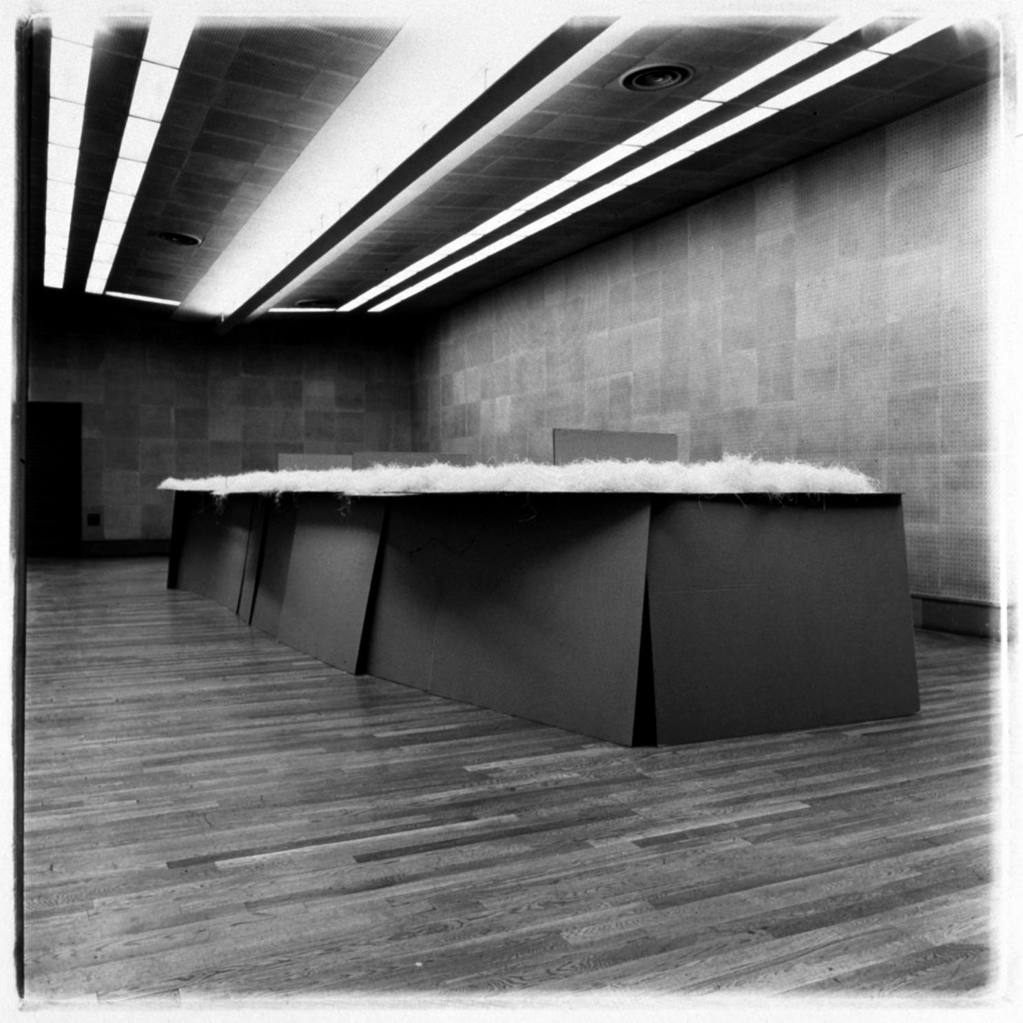 Installation photograph. Black and white photograph shows a long, table-like sculpture topped with what looks like light-coloured, fleecy material. Black panels lean against each other to make a base. The gallery has wood floors and textured walls.