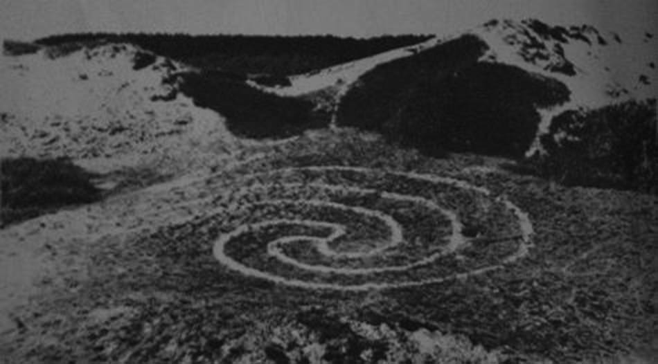Black and white photograph. A spiral swirls in light lines against a darker, grassy landscape.