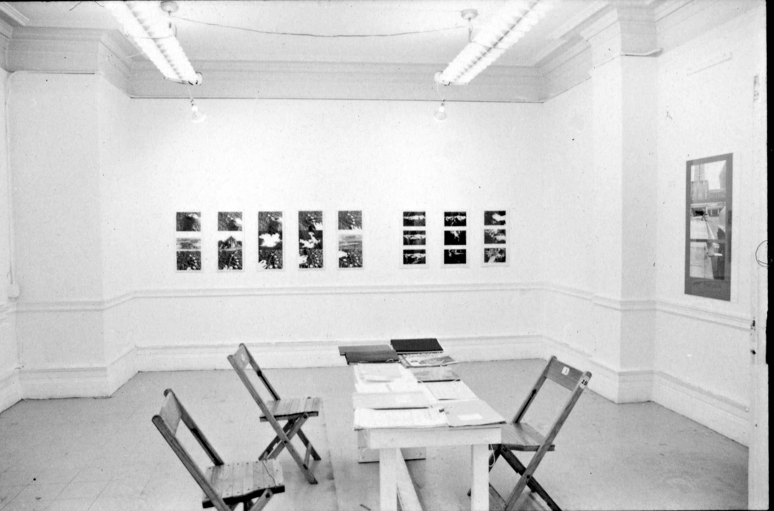 Installation photograph. Black and white photograph of three folding chairs sitting around a long table covered in piles of paper. Photographs and other works of art hang on the gallery walls.