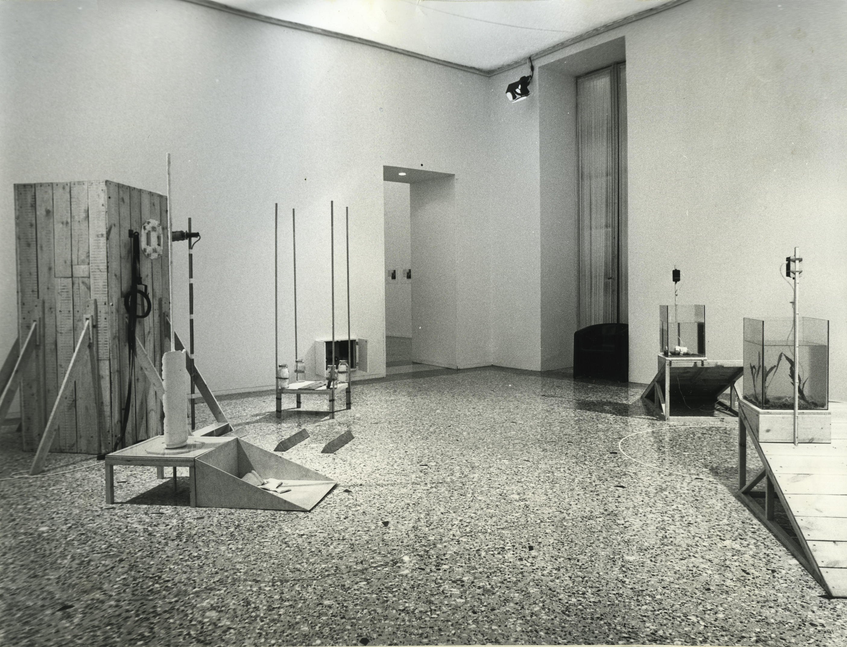 Installation photograph. Black and white photograph of a gallery space with five structures. Each of two fish tanks sit at ramps going down in opposite directions. A cabinet-like structure is across the room.