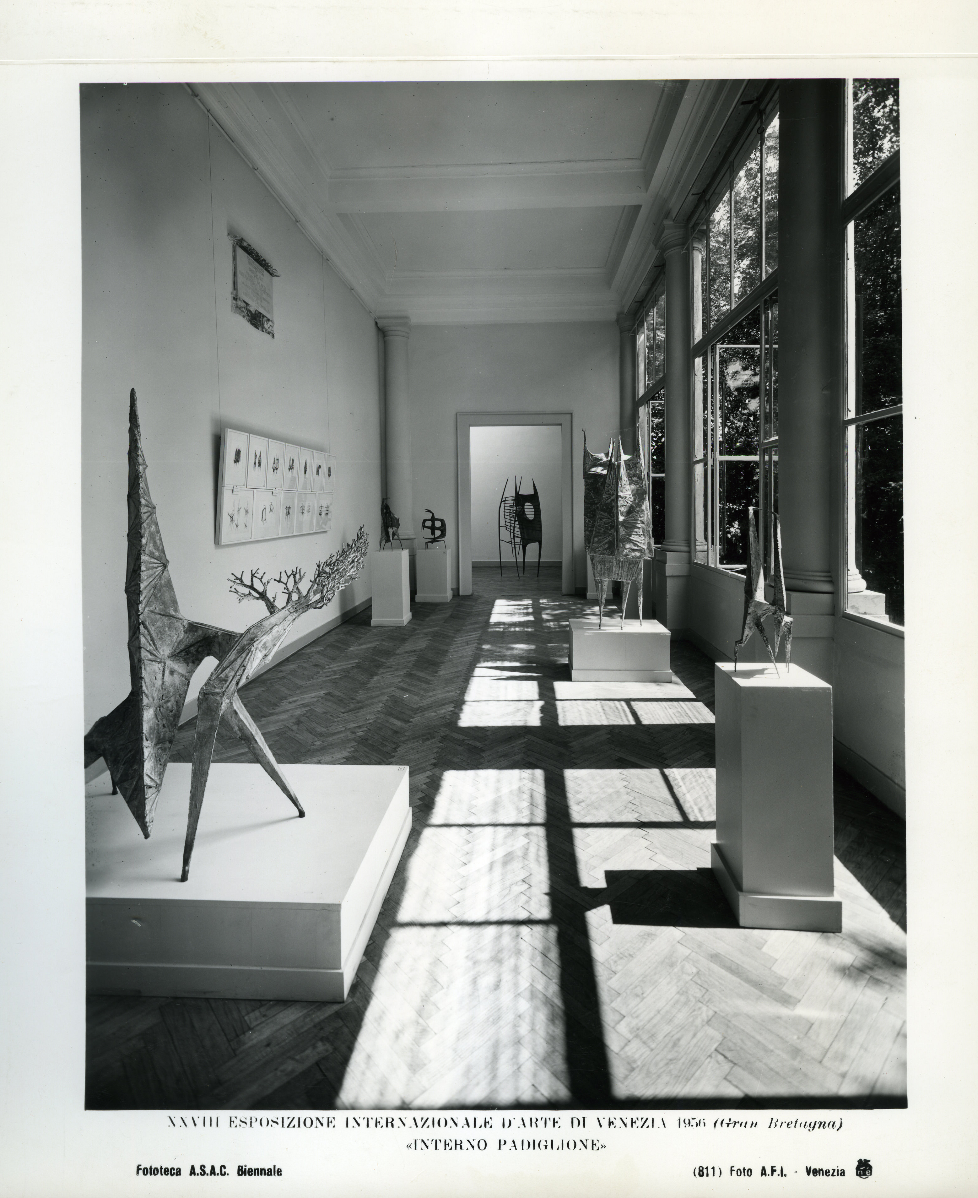 Installation photograph. Black and white photograph of a long, hall-like room lined with windows to our right. Sculptures are set out on plinths along the space, and a multi-piece work is hung on the wall to our left.
