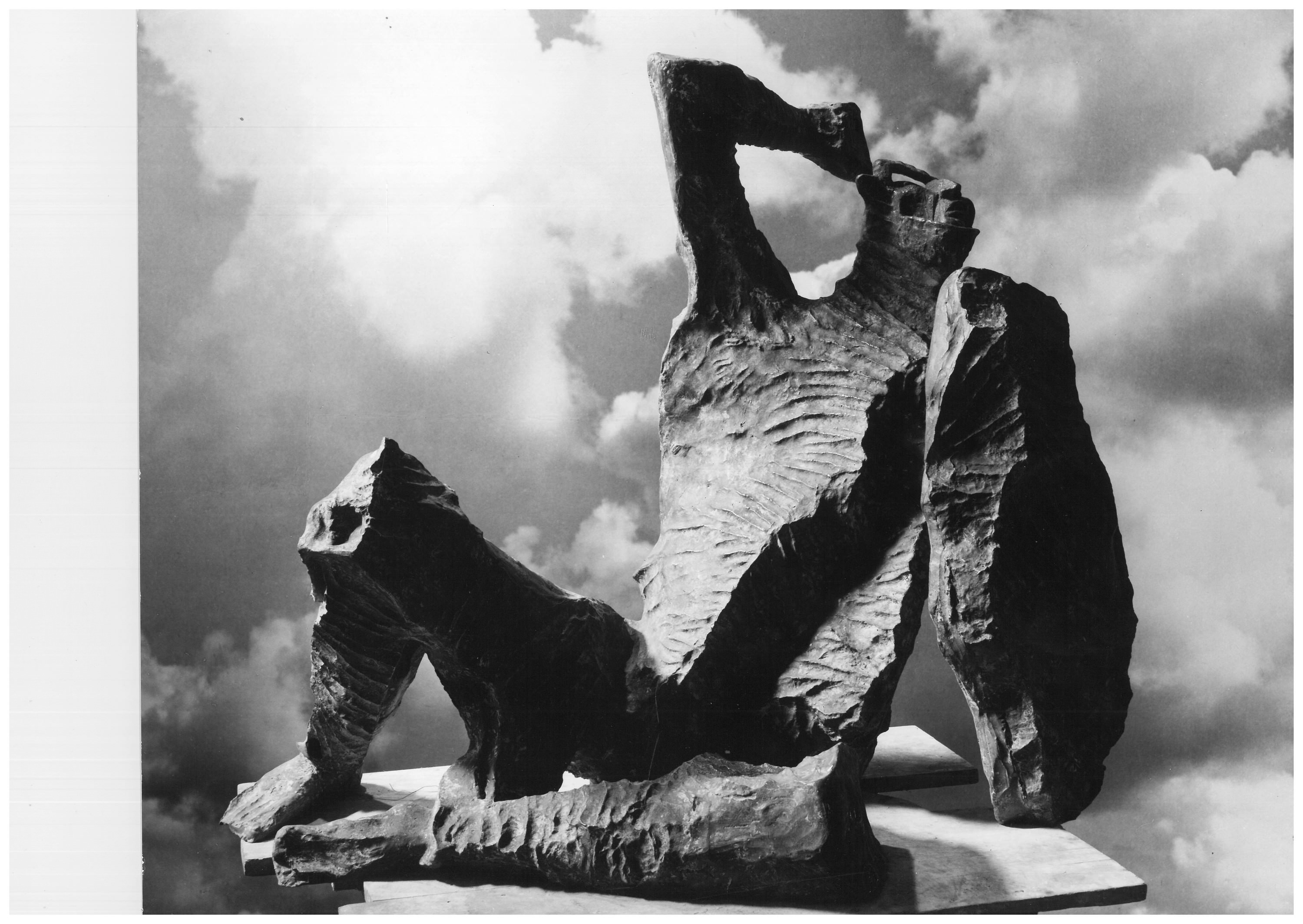 Sculpture. Black and white photograph of a freestanding, bronze, abstracted human form half reclining with a shield in one hand and the other raised to the head. The surface of the bronze is rough as if extra material had been chiselled off.
