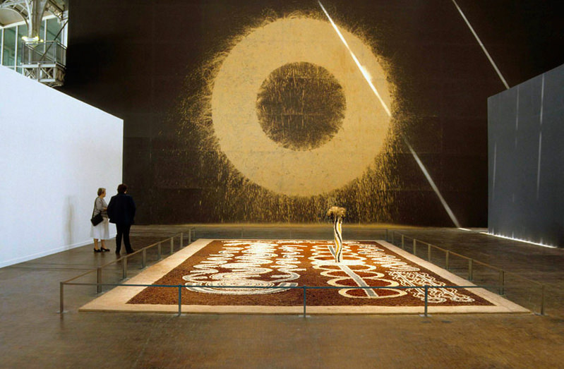 Installation photograph. A gold ring on a black wall is about two storeys high. The inner and outer edges are spattered, giving it a fuzzy look. A rectangular piece on the floor stretches between us. The floor piece has circular and wavy patterns in white against a deep red field. Two figures stand to our left observing the gold ring.