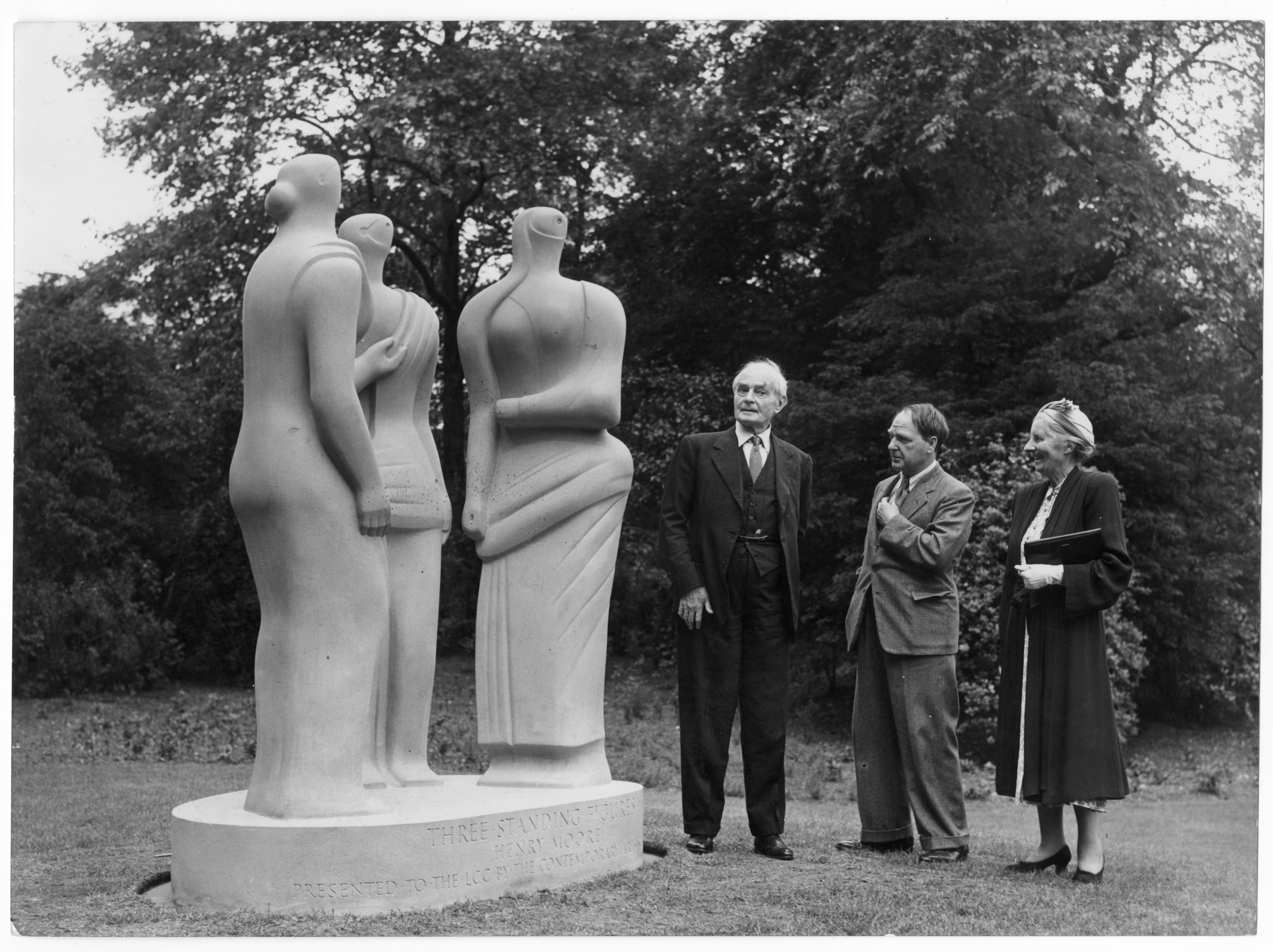 Black and white photograph. Three people stand in a park-like setting next to a light stone sculpture. It has an abstract style and shows three figures with simplified features. The people come up to about elbow height against the sculpted figures.