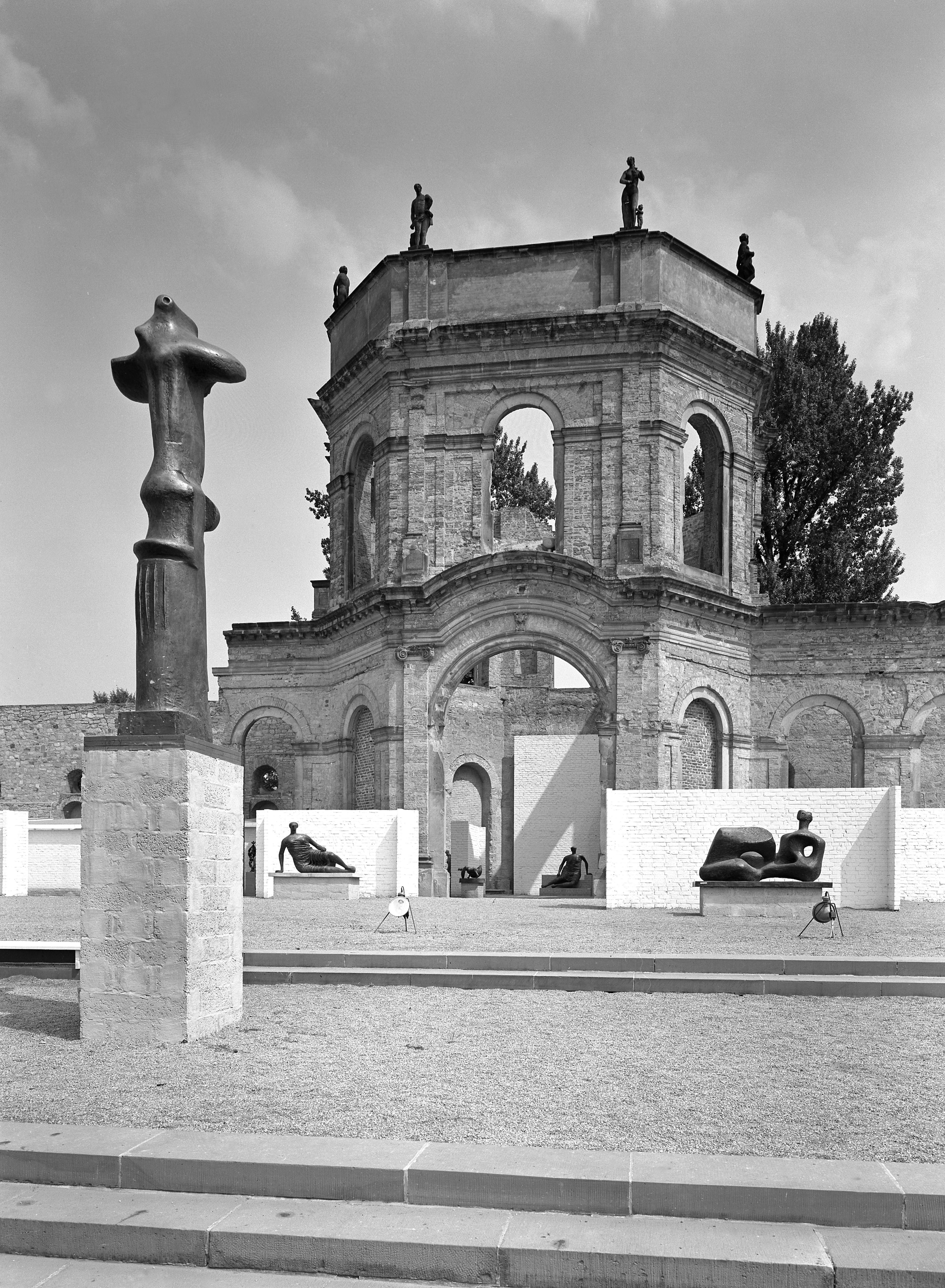 Installation photograph. Black and white photograph shows sculptures displayed against white walls set up in front of the remains of a stone building. Most of the sculptures are on plinths in this view, and the few near the central, arched doorway of the building are of stylized human bodies.