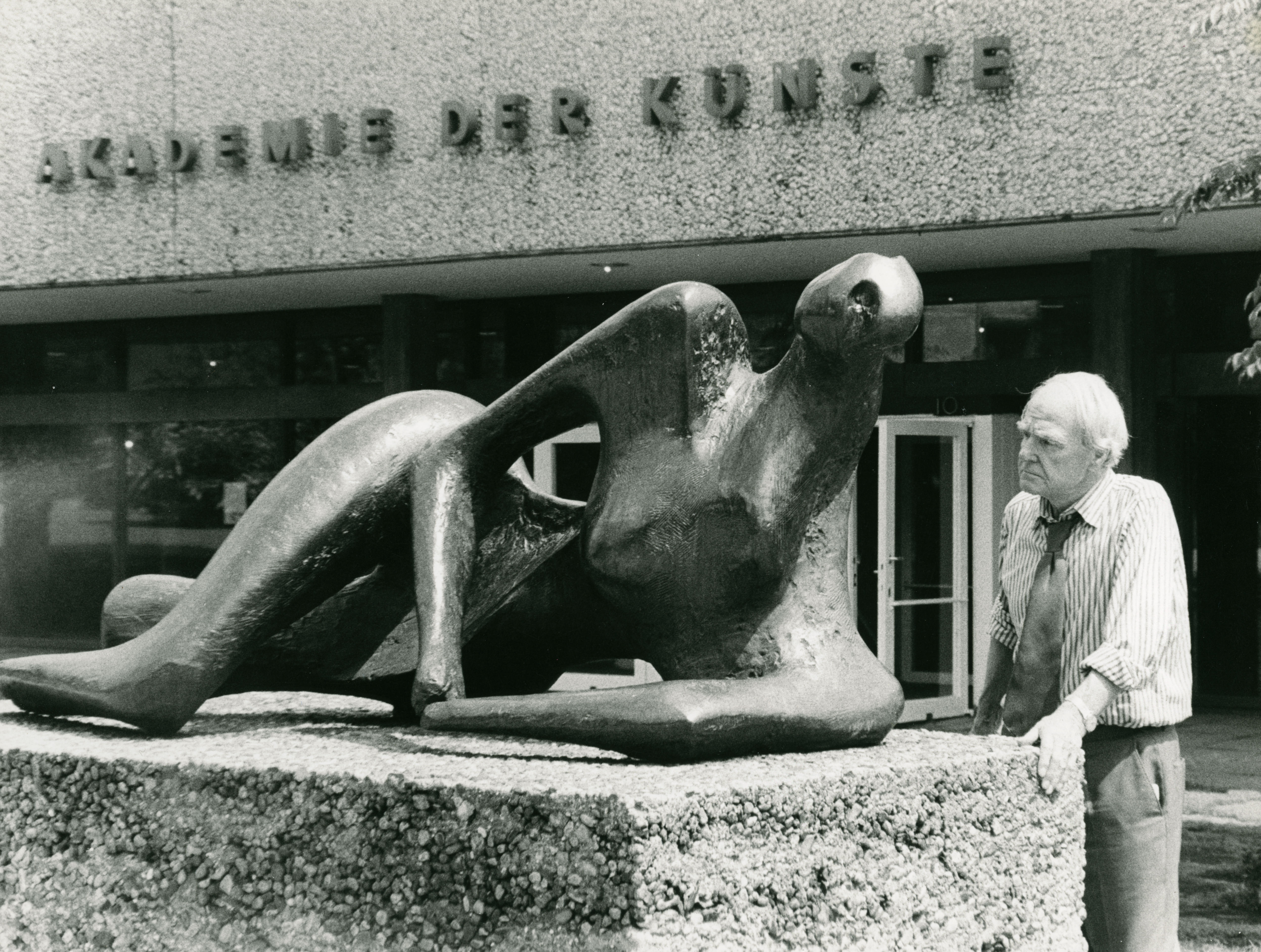 Black and white photograph. A white-haired man with a wrinkled face, Henry Moore, looks under lowered brows at a bronze sculpture of on a waist-high plinth. The sculpture is a stylized reclining female form. The building behind him has the sign, “Akademie der Kunste”.