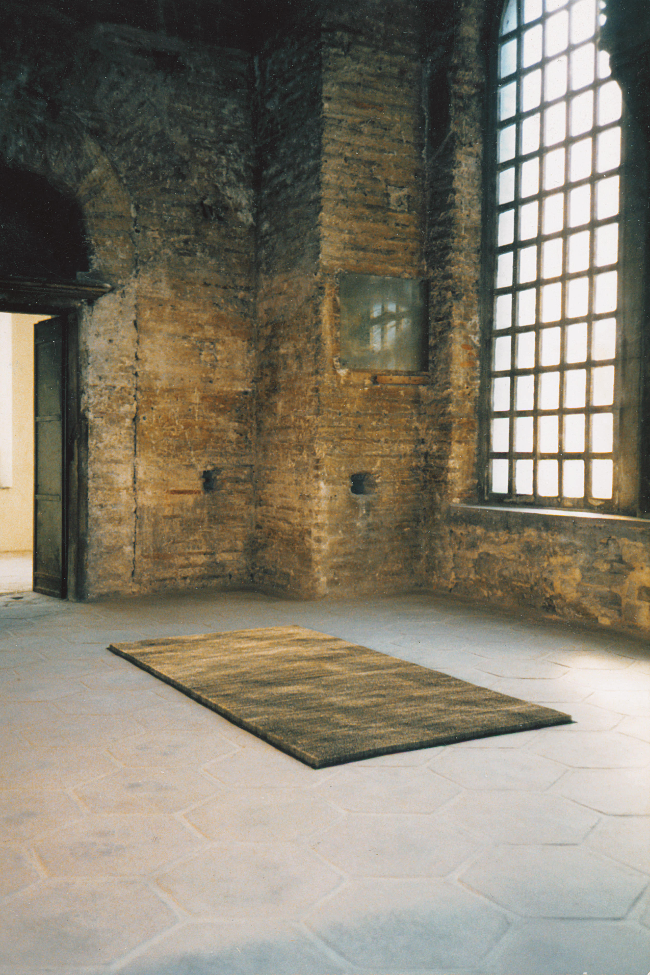Sculpture. A rectangle the size of an area rug sits on the stone floor of a stone room. The sculpture is streaked with golden-brown and black. From this distance, it appears to be a carpet. Close up, we find it is made from rows of pins.