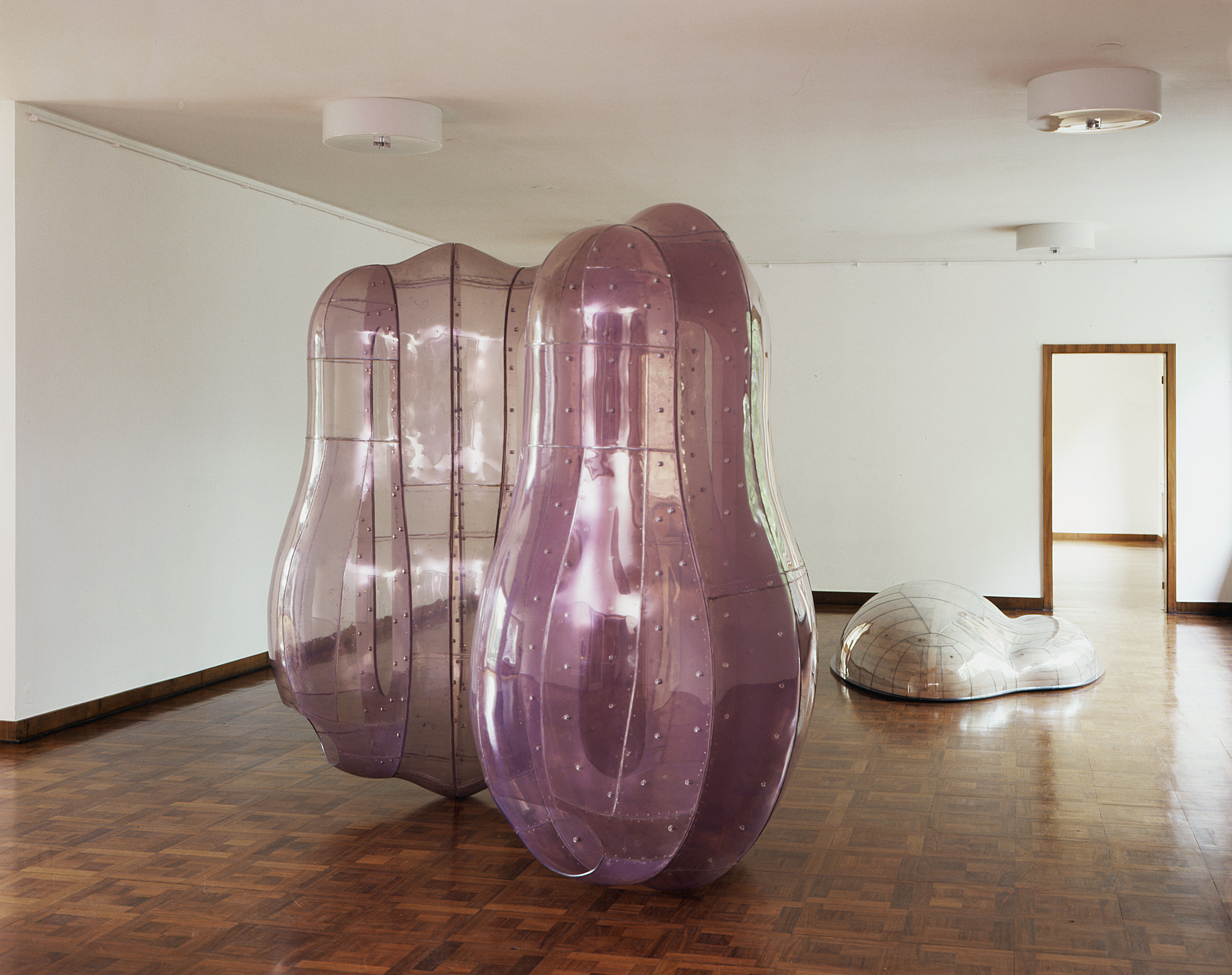 Installation photograph.Three abstract sculptures are displayed in a gallery with white walls and parquet floors. Two forms close to us are transparent pale purple. Shaped like widened bowling pins, they nearly reach the ceiling. A similar form in white lies on the floor.