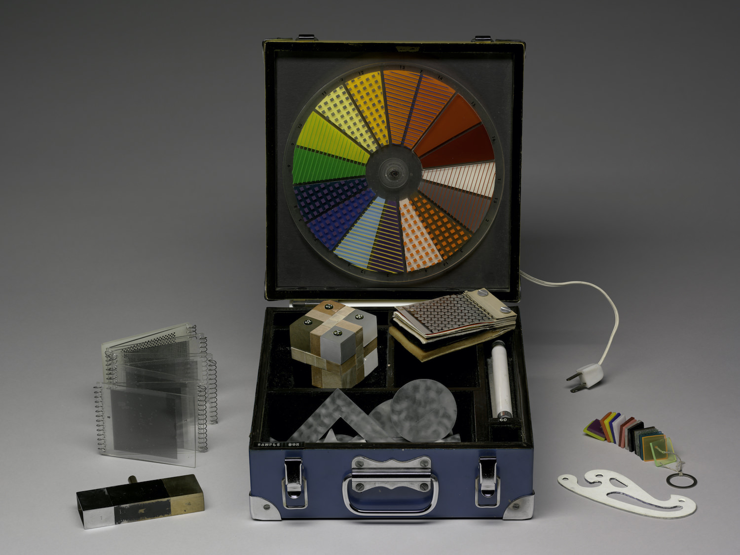 Sculpture. A blue case holds samples of materials and a neon light in compartments in the bottom. A Plexiglas wheel with slices of colour and pattern is mounted to the open lid like a turntable. Other materials from the sample box are displayed around it.