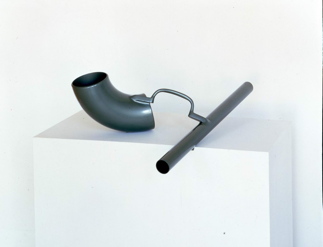 Sculpture. Two segments of tubes are attached with a handle-shaped piece of metal. The tube to our left is wide, short, and curved, like macaroni. The tube to our right is narrow and long, and it rests in a notch cut out in the front edge of the plinth.