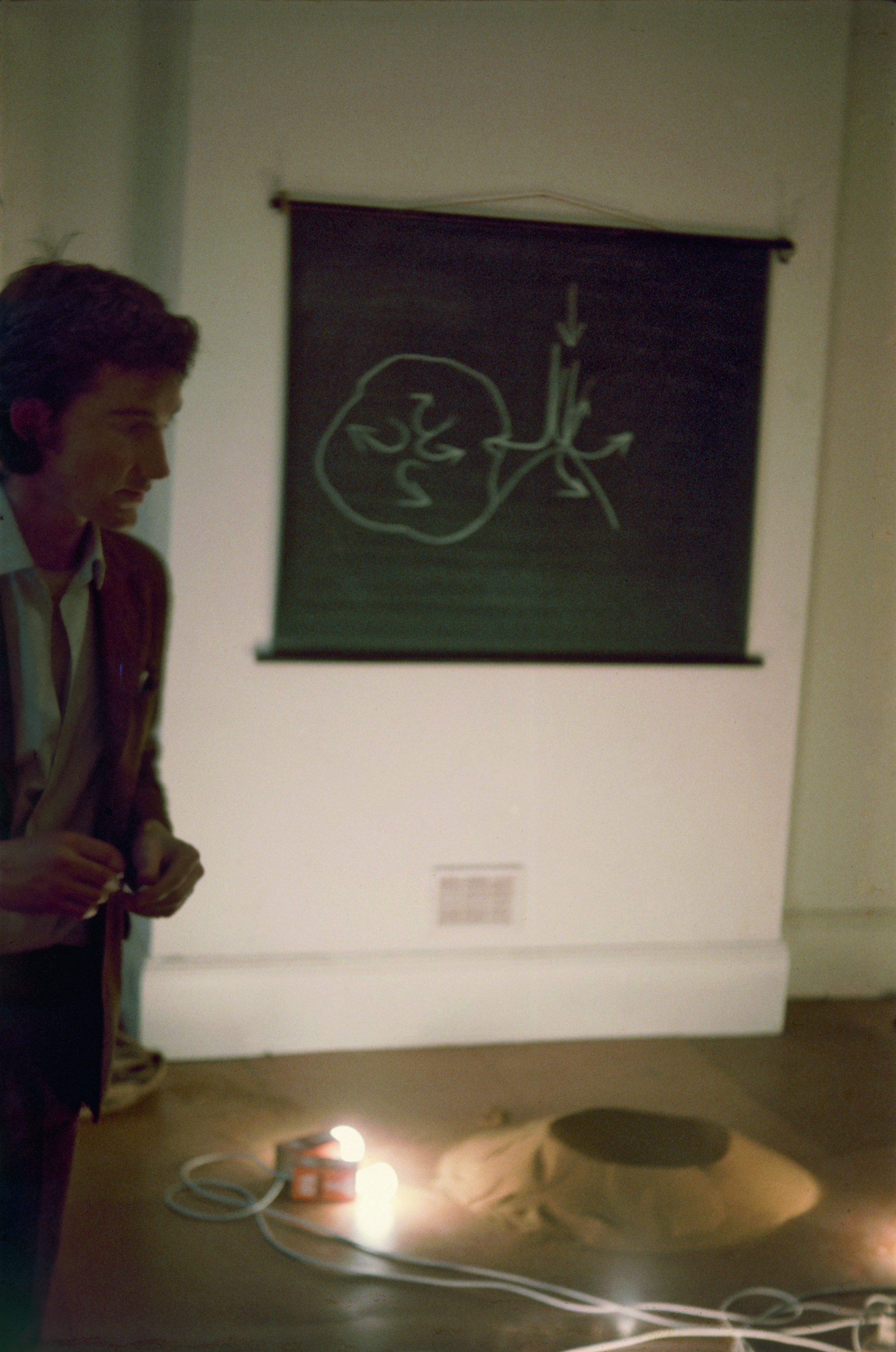 Colour photograph. A pale-skinned man with dark hair stands to our left, almost out of frame. A black picture with a drawing or diagram in white hangs on the wall behind him. Two lights lie on the floor next to a tan-coloured mound.