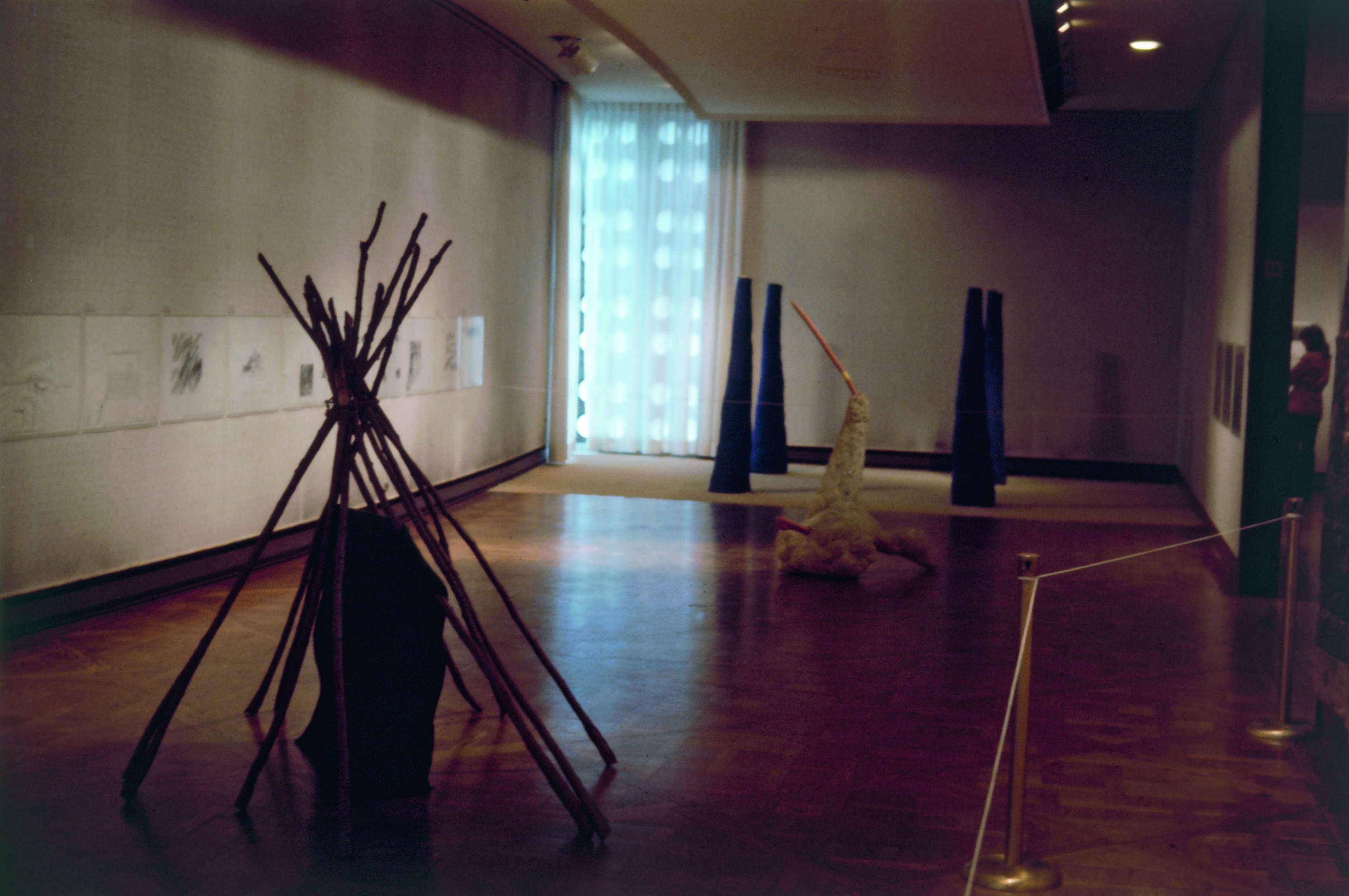 Installation photograph. Sculptural pieces made from wood and other material sit on the floor of a dim gallery. Works of art line the walls.