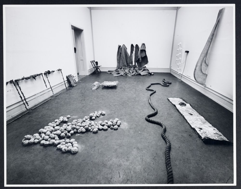 Installation photograph. Black and white photograph of a gallery space. Several abstract sculptures lie on the floor. Another leans against one wall, two are affixed to the opposite wall, and the piece farthest from us appears to be made of cloth hung on the wall and pooling on the floor.