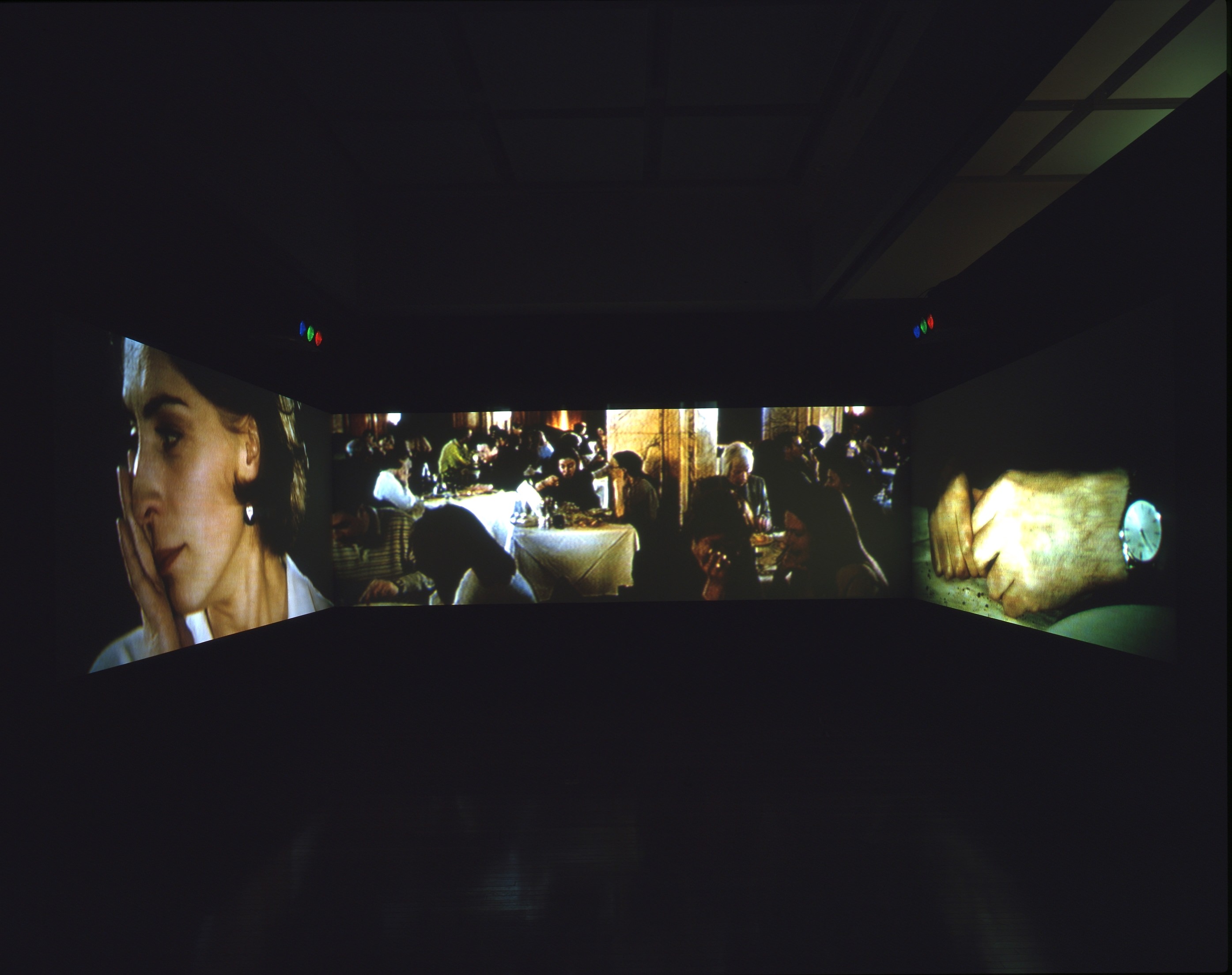 Installation photograph. Three screens wrap around the walls in front of us in a dark room. A woman touches her face on the screen to our left. People gather in a restaurant on the middle screen, and the third shows a close-up of a pair of hands.