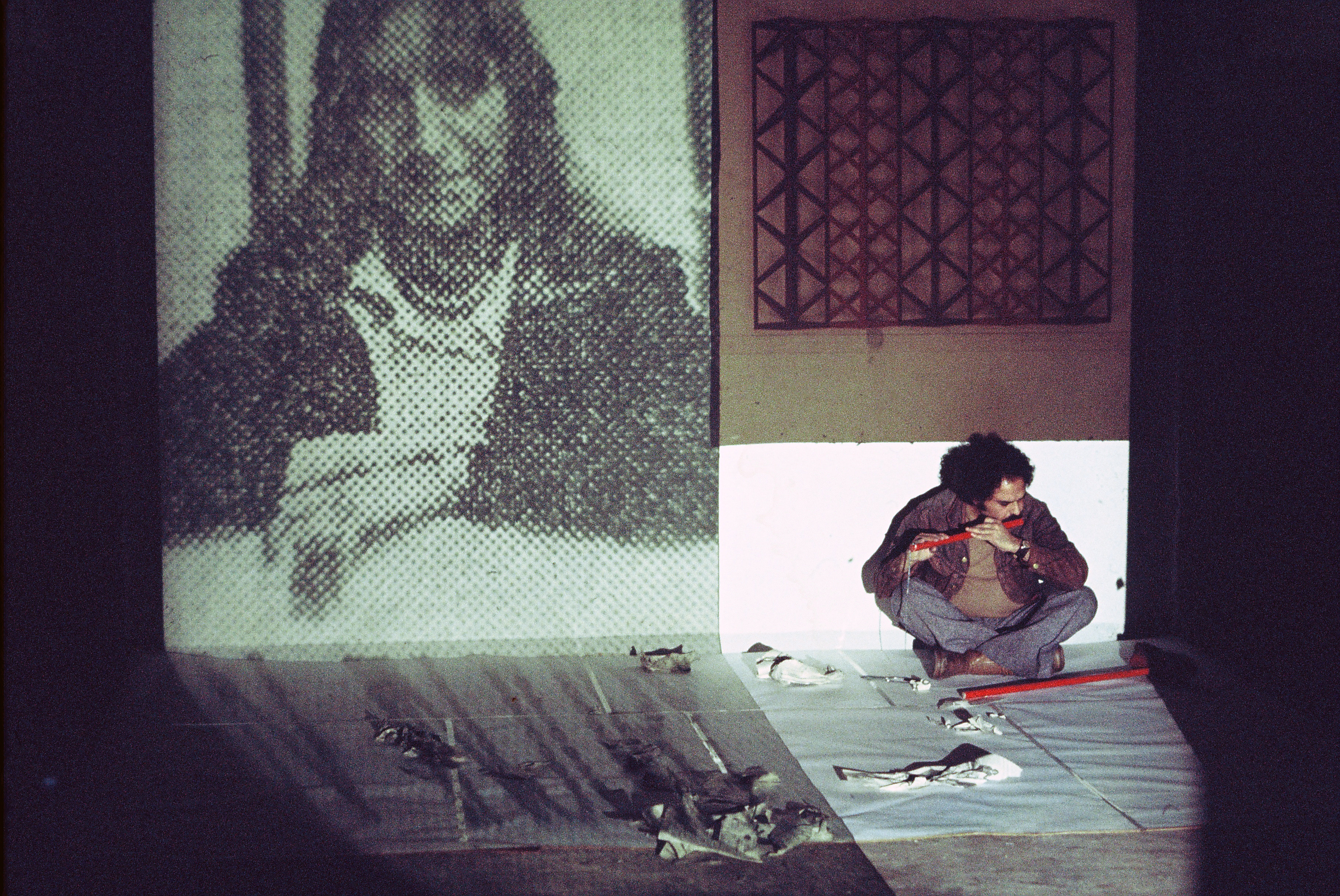 Performance art. A shaft of light falls on a brown-skinned, moustached man sitting cross-legged against a wall as he plays a red flute. A black and white photograph of a person with long hair is on the wall to our left, benday dots visible. Tools or other materials are laid in front of and beside the man.