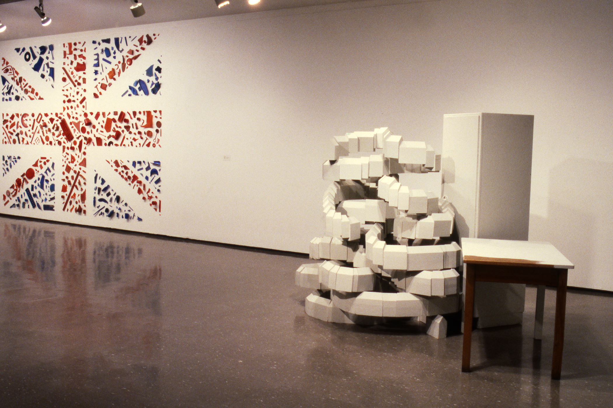 Installation photograph. A Union Jack spans the height of a gallery wall to our left. The red stripes and blue triangles are made from found objects. A work to our right looks like white blocks are stacked in such a way that long rows spiral upwards in a way that defies logic.