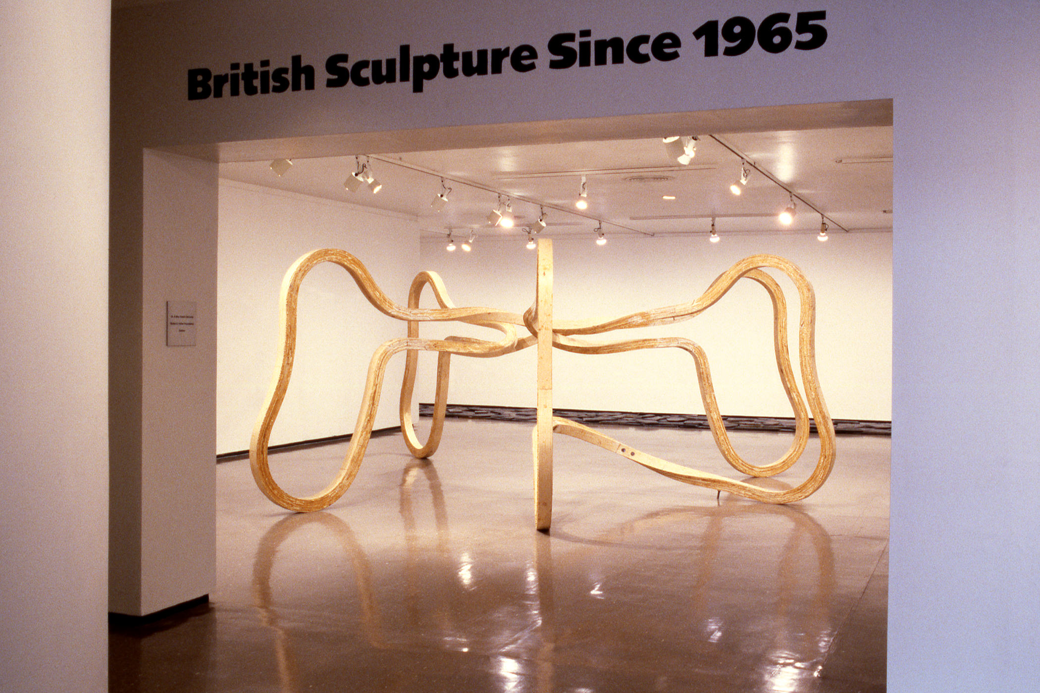Installation photograph. In the gallery beyond and framed by the opening, an abstract sculpture made from bent wood curves out and down from a central point.