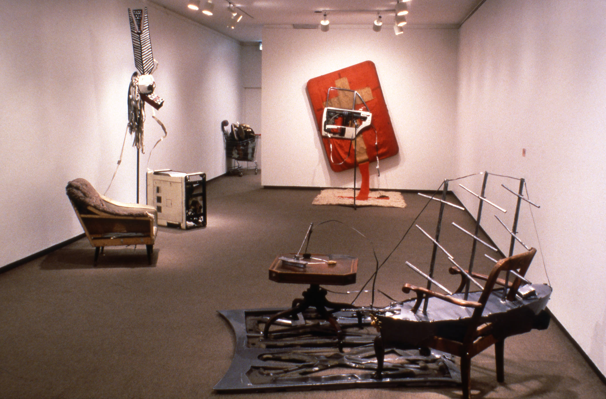Installation photograph. Six sculptures incorporate some familiar objects, like an armchair next to an old-fashioned washing machine and a shopping cart in the next room. Some are surreal, like a chair whose seat has been replaced with a model of a boat. Another is abstract.