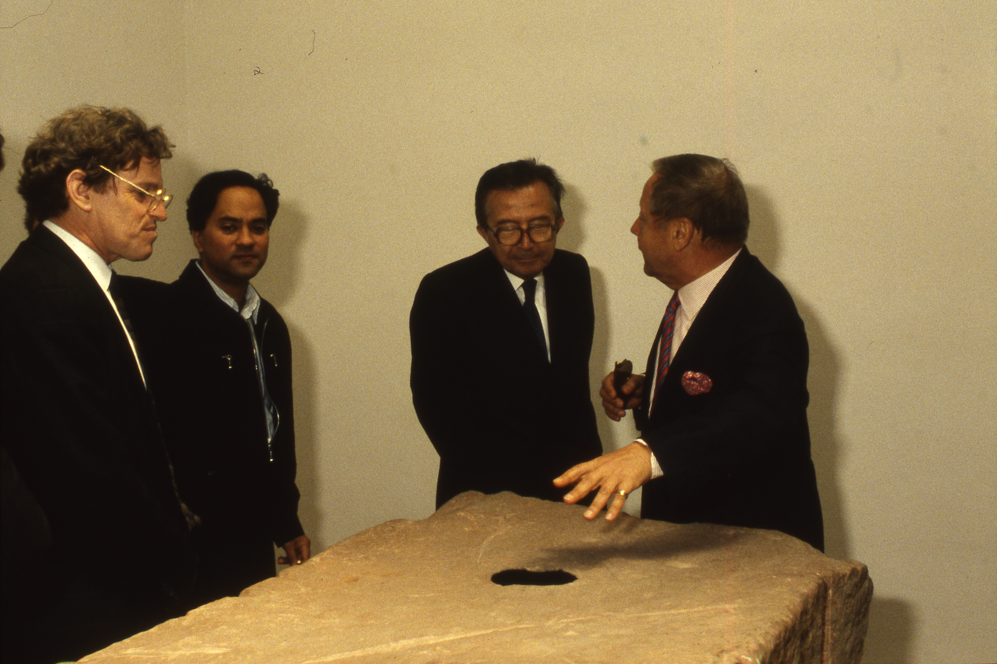 Colour photograph. Four men gather around a roughly rectangular, red sandstone block. The man to our right gestures at a black circle on the top.