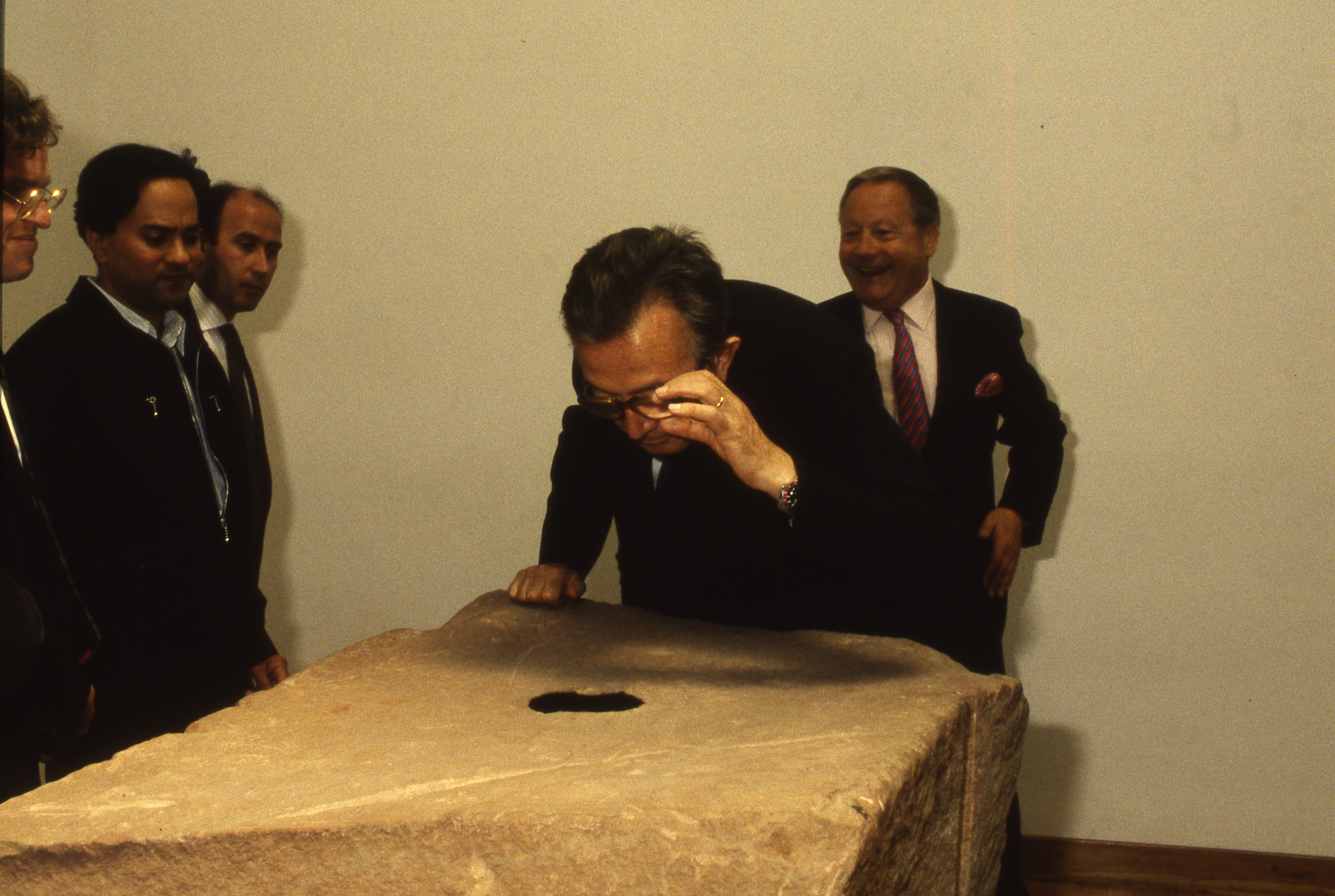 Colour photograph. Five men gather around a roughly rectangular, red sandstone block. One man braces himself with a hand on the edge of the block and leans forward to look at a black circle on the top. He adjusts his glasses with the other hand.