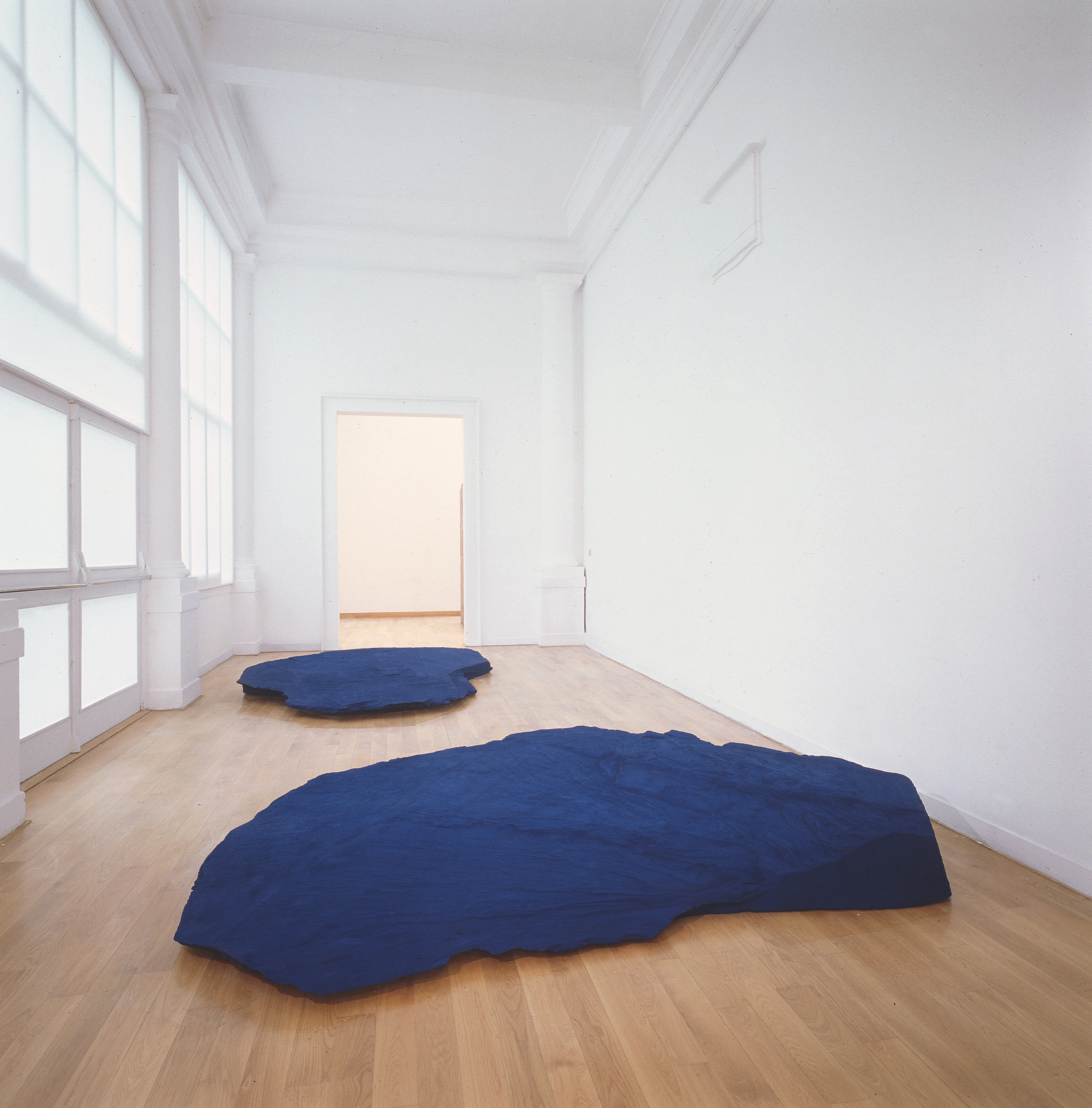 Installation photograph. Two pieces of slate sit on the floor of a hall-like room lined with windows to our left. The slate is painted royal blue and are placed such that a visitor would need to snake through the room between the works.