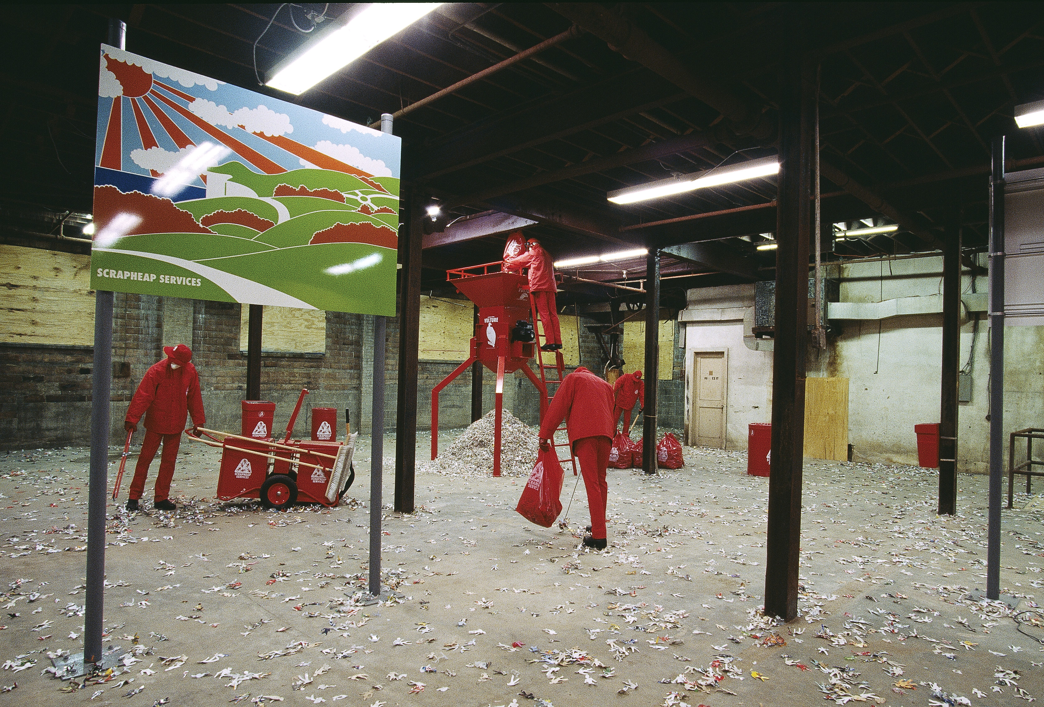 Installation photograph. Five mannequins wearing all red, pick up litter with grabbers, though the whole scene is set in a room with I-beams holding up exposed ceilings. One mannequin dumps a bag into a shredder. A sign near us reads “Scrapheap Services” under a cartoonish landscape.