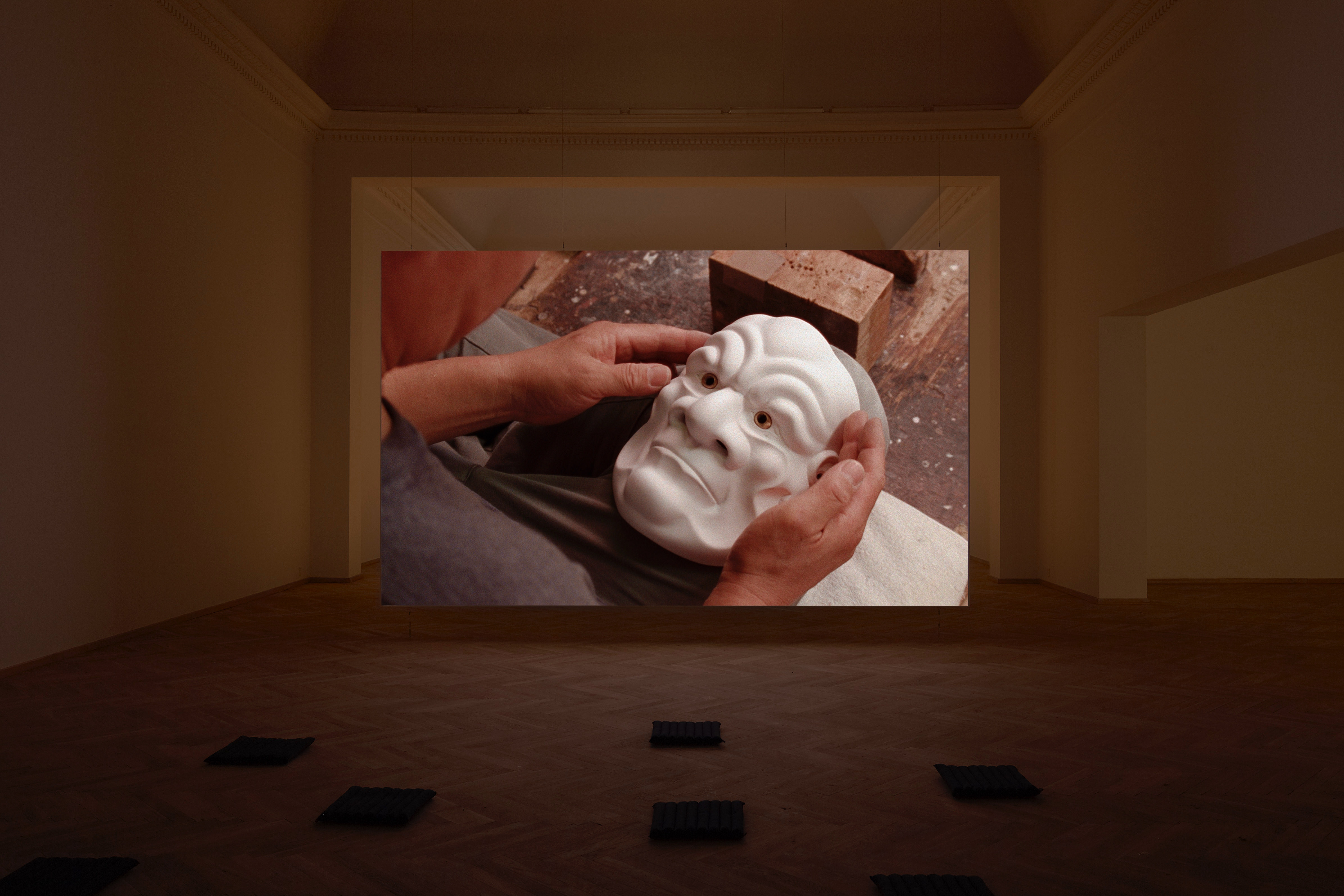 Installation photograph. A film is projected at the end of a dim gallery. The still shows hands holding a mask of a face with exaggerated wrinkles. The face’s surface is white.
