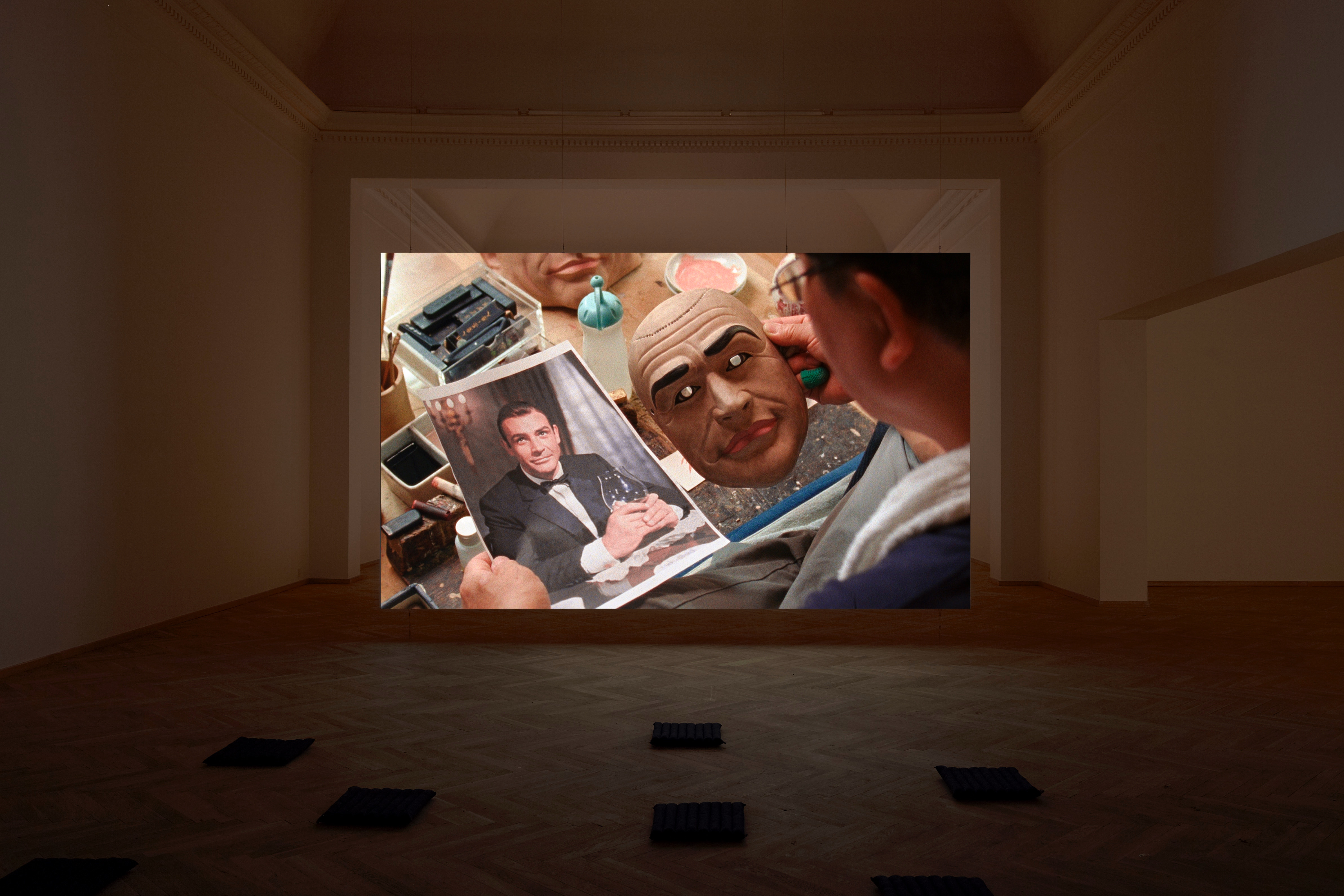 Installation photograph. A film is projected at the far end of a dim gallery. In it, a person holds a mask in one hand and a photograph, both of Sean Connery, in the other.