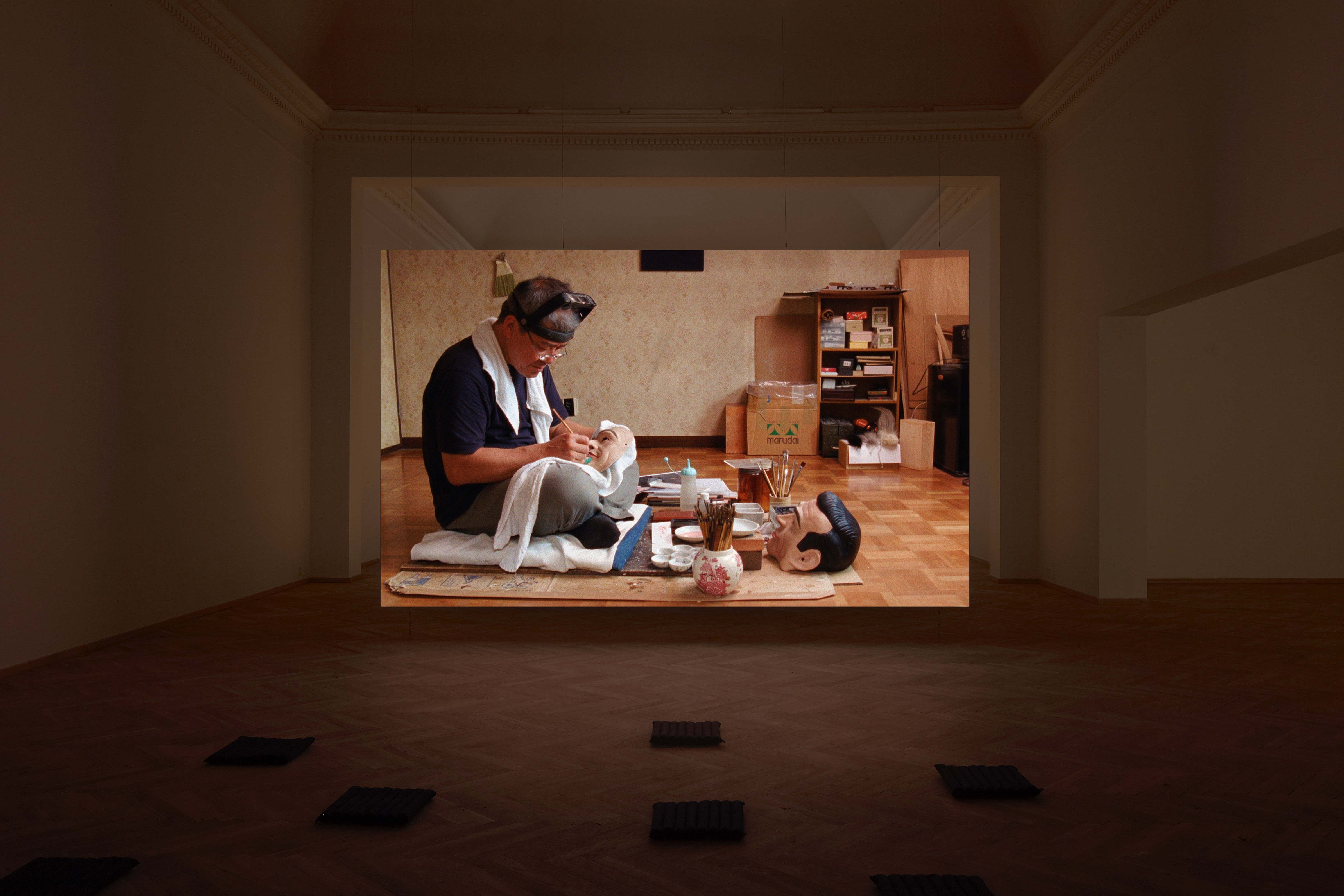 Installation photograph. A film is projected at the far end of a dim gallery. In it, a man sits cross-legged on the ground, painting a mask. Paint brushes and paint are arranged on a mat in front of him.