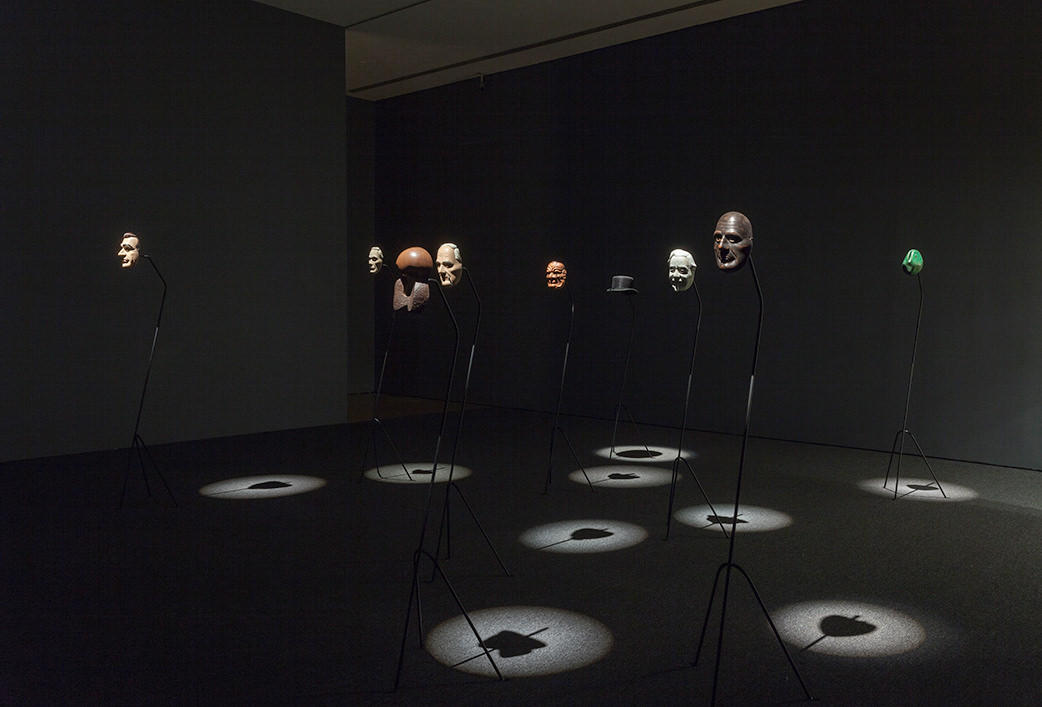 Film still. Five masks, a head-sized abstract sculpture, a top hat, and a green form are on stands in a darkened gallery. Each is spotlit from above.