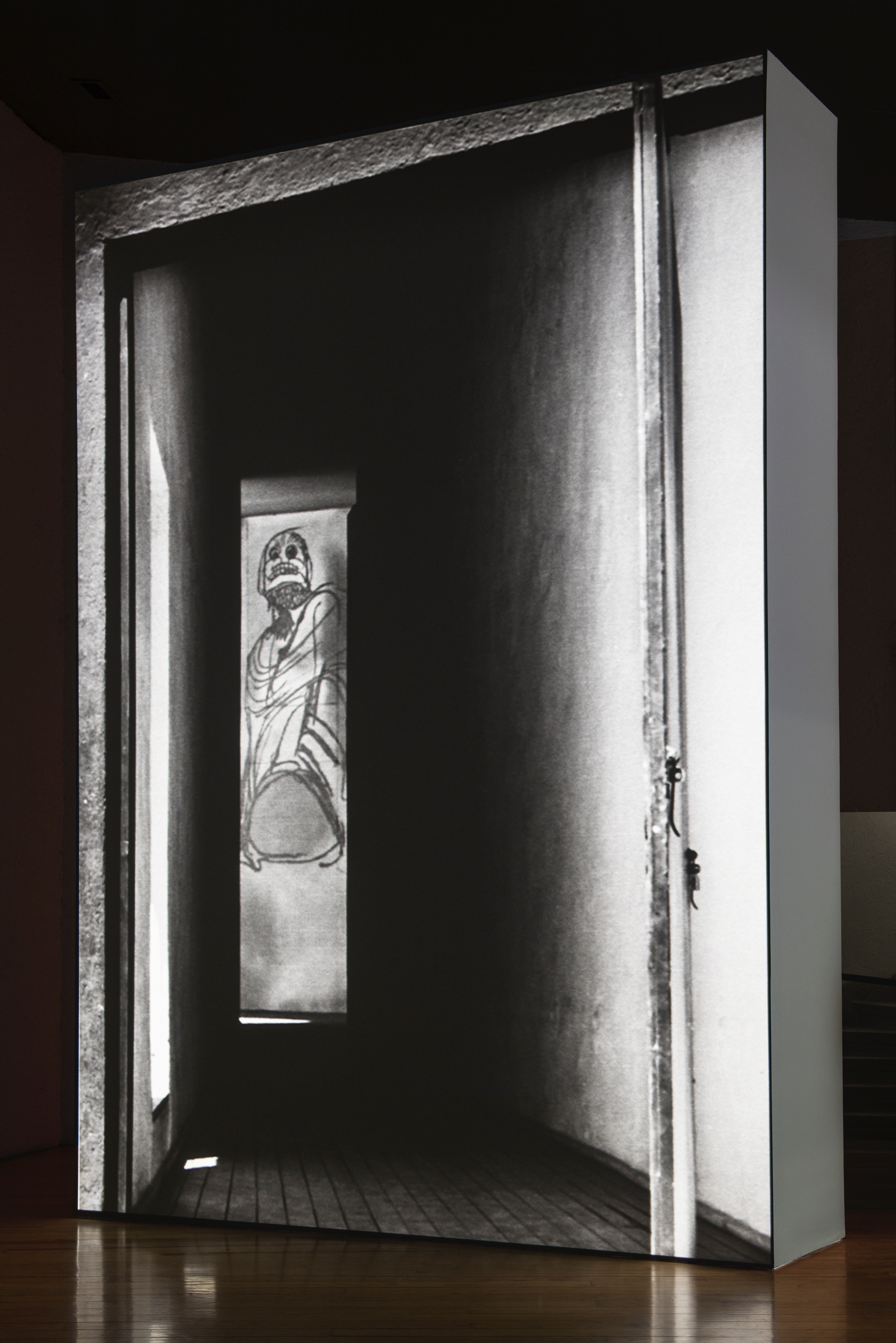 Installation photograph. A black and white film is projected onto a floating wall. This still shows a tall, narrow drawing seen at the end of a hall. The drawing is of a stylized person with a skull-like face.
