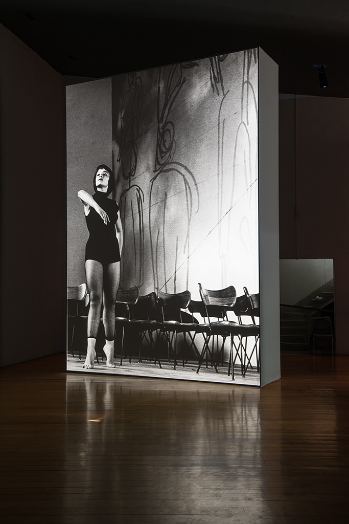 Installation photograph. A black and white film is projected onto a floating wall. The still shows a woman with dark hair wearing a black leotard and standing on tiptoes next to rows of wooden chairs. One elbow is gracefully lifted, mid-dance.