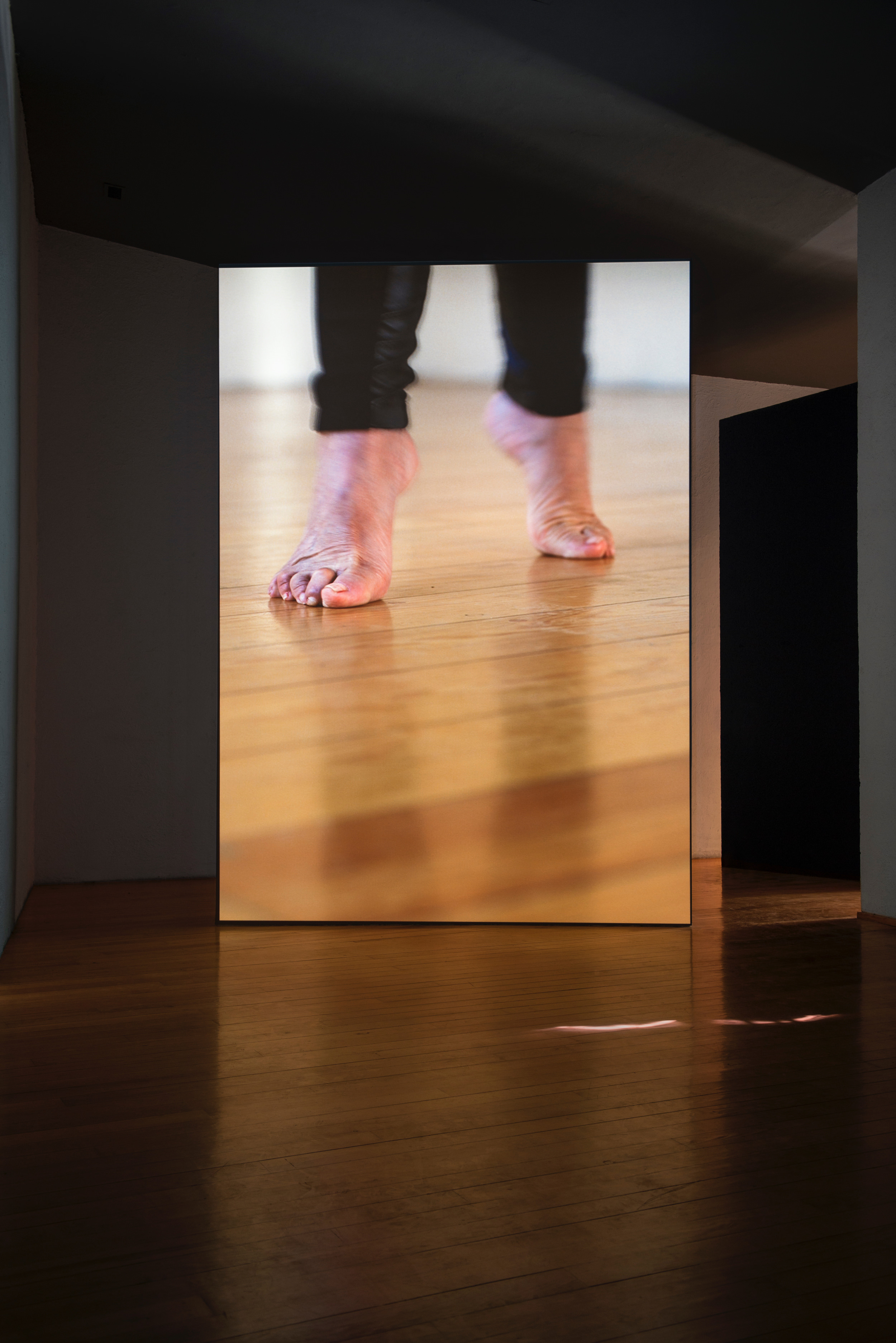 Installation photograph. A film is projected onto a floating wall. This still shows a close up of aged, bare feet, lifted on tiptoe on a wood floor.