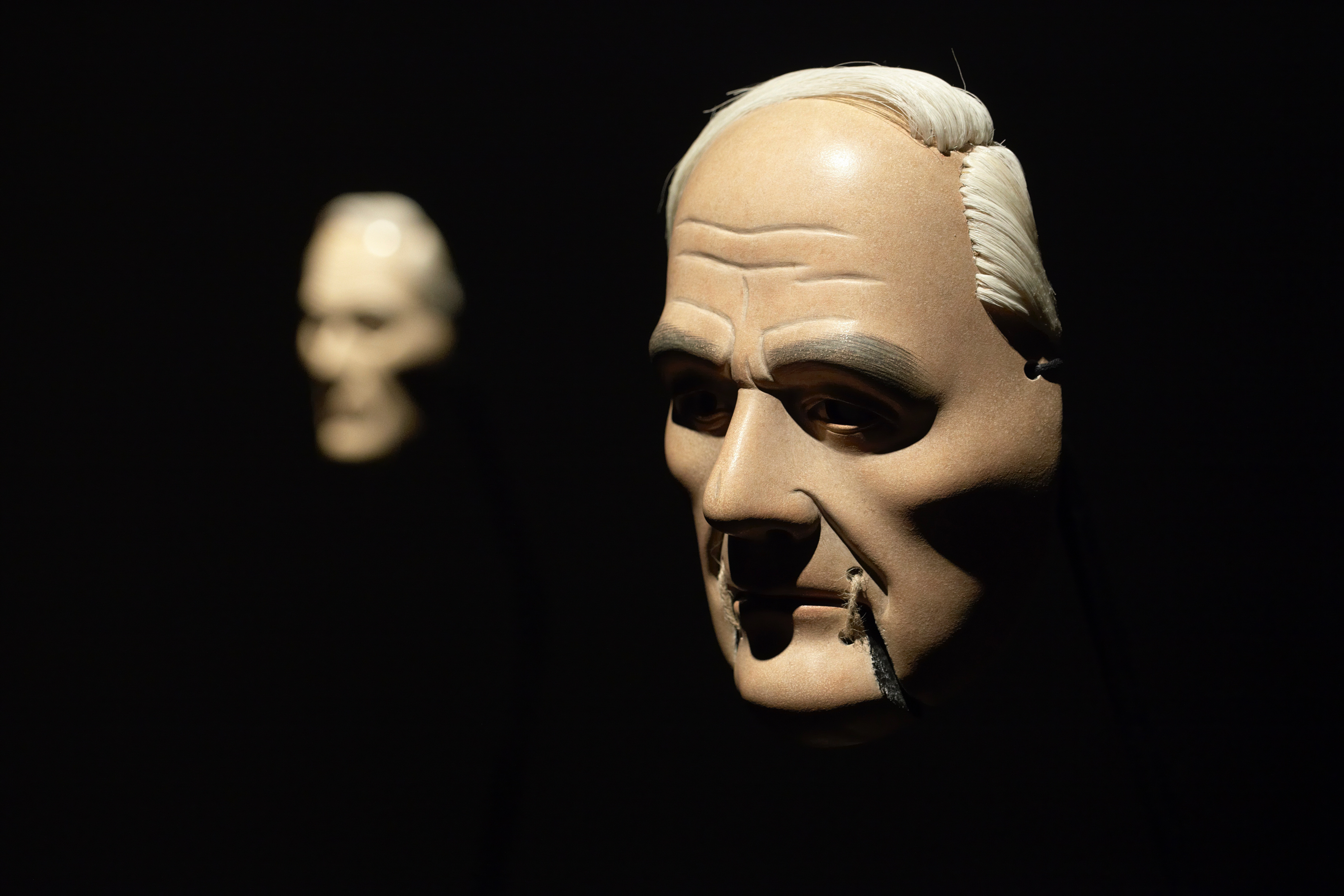 Film still. Two masks float against an ink-black background. The one closer to us has pale skin, white hair, high cheekbones, and the chin is tied to the mouth with twine. The other mask is similar but out of focus in the background.