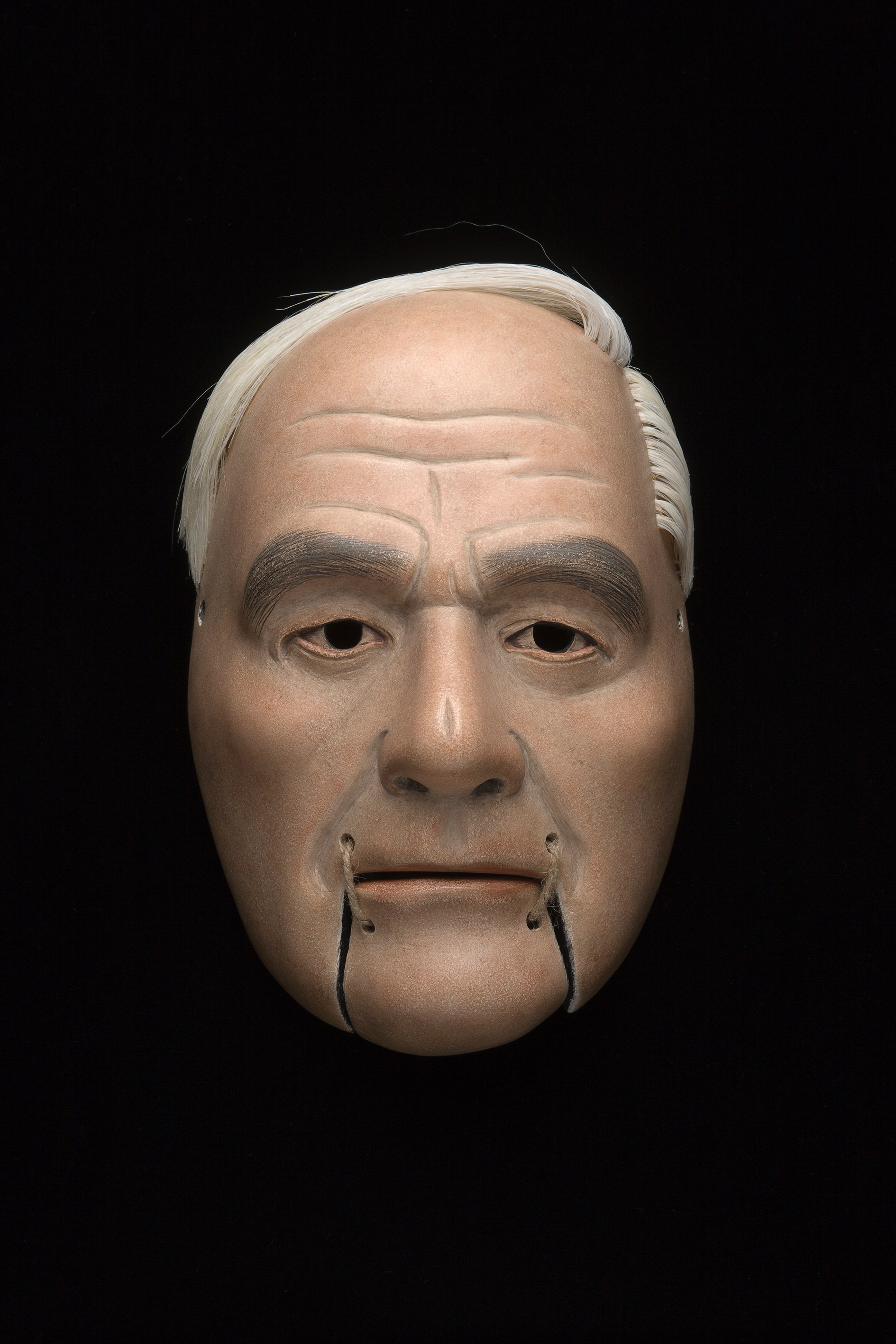 Sculpture. Wooden mask of a man’s face has pale skin, white hair, and wrinkles across the forehead. Hooded eyes look out under heavy black brows. The moveable chin is tied onto the mouth with twine.