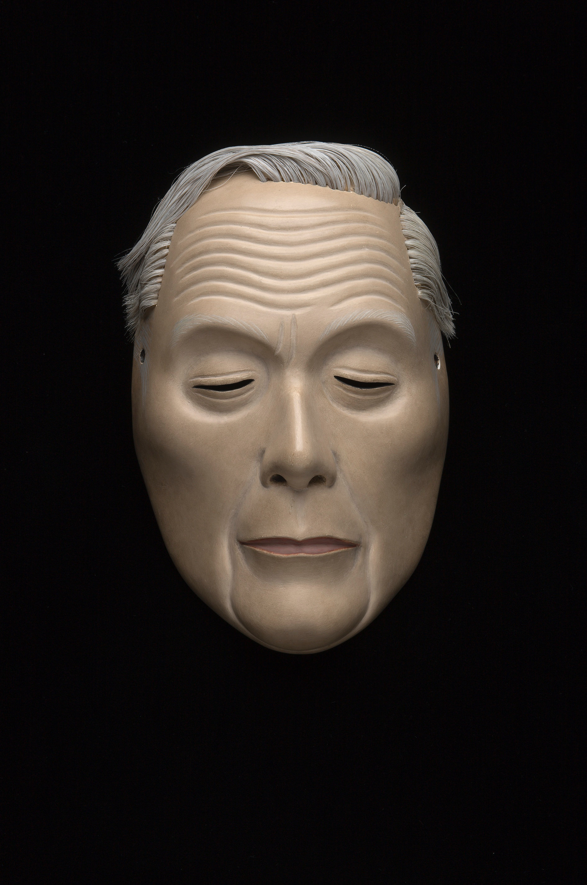 Sculpture. Wooden mask of a man’s face has short, grey hair, a ridged forehead over raised, arched brows. The downcast eyes are curved slits. The man’s thin lips are set in his oval face.