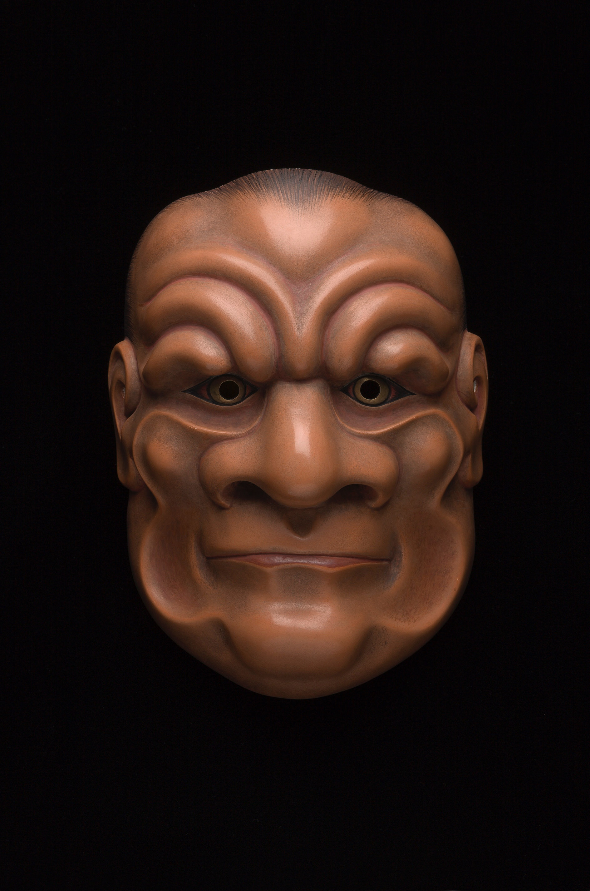 Sculpture. Wooden mask of a stylized man’s face. Wrinkles curving from the wide nose up over eyes and those lining the cheeks are exaggerated. The upper lip disappears over a thin lower lip. The skin is light brown.