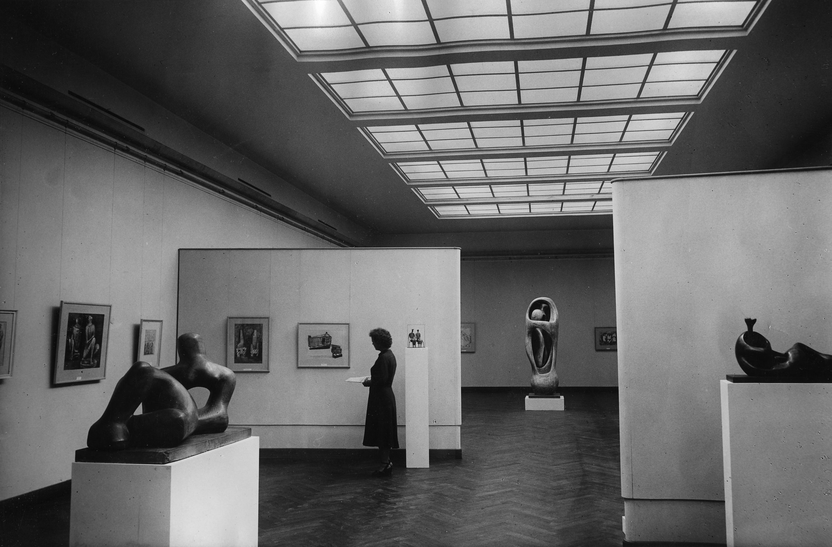 Installation photograph. Black and white photograph of sculptures of abstracted bodies displayed on plinths in a gallery hung with works of art.