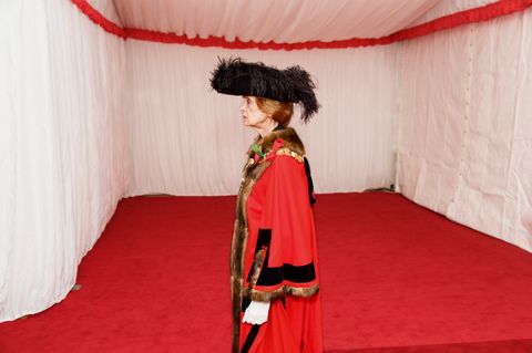 Colour photograph. Shown from the knees up, a woman wearing red regalia trimmed in black, gold, and fur faces our left in profile. Her black hat is lined with feathers, and the white-gloved hand we can see is in a loose fist at her side. She stands in what appears to be a white tent.