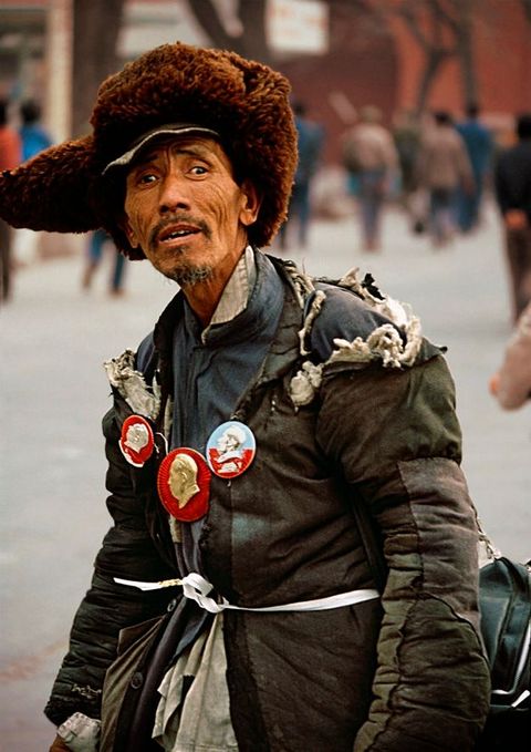 Man with medium brown skin looks at us on a city street. He wears a fuzzy brown hat with ear flaps and two winter coats tied around the waist with a white band. Three medallions or pins are affixed over his chest, each showing a person in profile. His eyebrows are gathered, and his hollow cheeks lined.