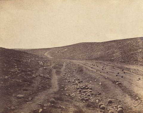Black and white photograph. Dozens of cannonballs are strewn along a road and ditch in a shallow, barren valley.
