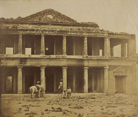 Sepia-toned photograph photograph. Four dark-skinned men and a white horse stand in front of the ruins of a two-storeyed, colonnaded structure. Rubble is strewn along the street in front of us.